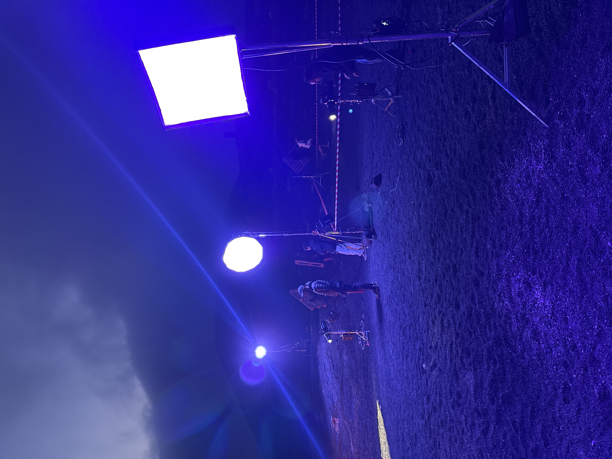 At night in Teguise, Spain, an outdoor film set is illuminated with three artificial light sources. At the top of the image is a square-shaped light panel on a stand. Further down is a spherical light on a stand. At the bottom of the image is another, smaller light source, also on a stand. Several people are on the set, including one crouched near the top light and another standing between the middle and bottom lights. There is film equipment visible, including a camera on a stand near the middle light. The environment is dark, with a grassy ground surface. Part of the sky and some clouds are visible in the lower-left corner.