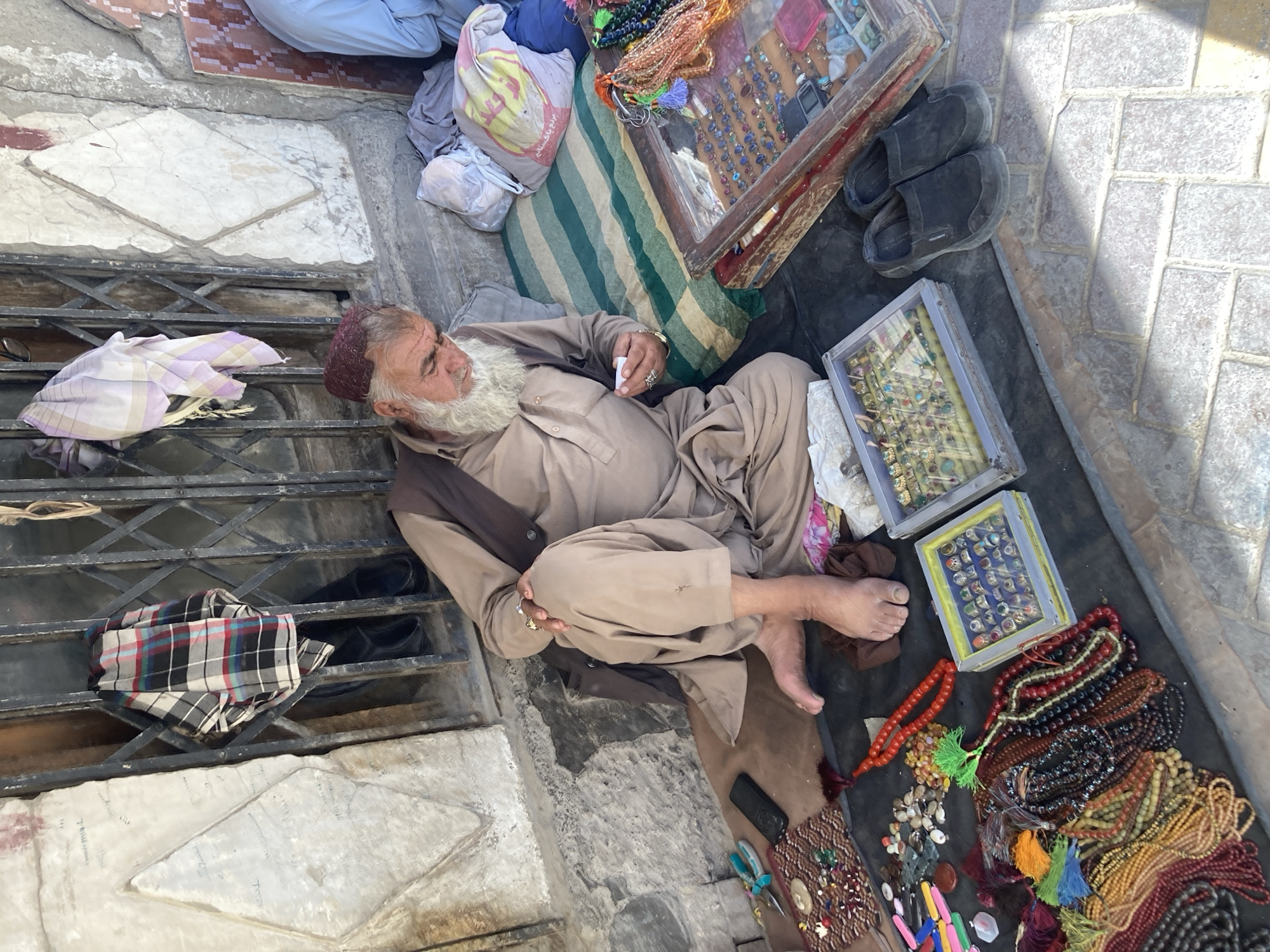 An elderly, bearded man wearing traditional Pakistani clothing rests on the ground next to a sidewalk grate, surrounded by his wares. These include jewelry boxes, rosaries, and other trinkets. The scene appears to be an outdoor marketplace in Quetta City Tehsil, Pakistan.