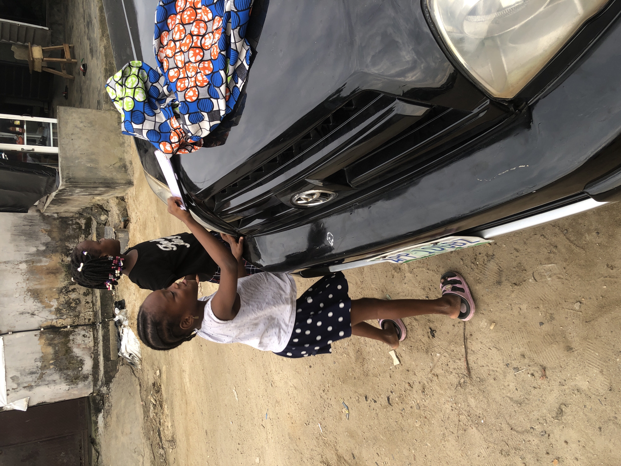 In Ado, Nigeria, two young girls are near a black SUV. The girl in the foreground, wearing a white top and a dark blue skirt with white polka dots, is reaching towards the hood of the car. A brightly colored fabric with circular patterns in blue, orange, and green is draped over the hood. Behind her, a second girl in a black t-shirt and dark hair braided with beads stands looking away. The car is parked on a dirt road next to a weathered concrete building. The environment appears to be an urban or semi-urban setting with sandy ground and a rough-textured wall in the background.