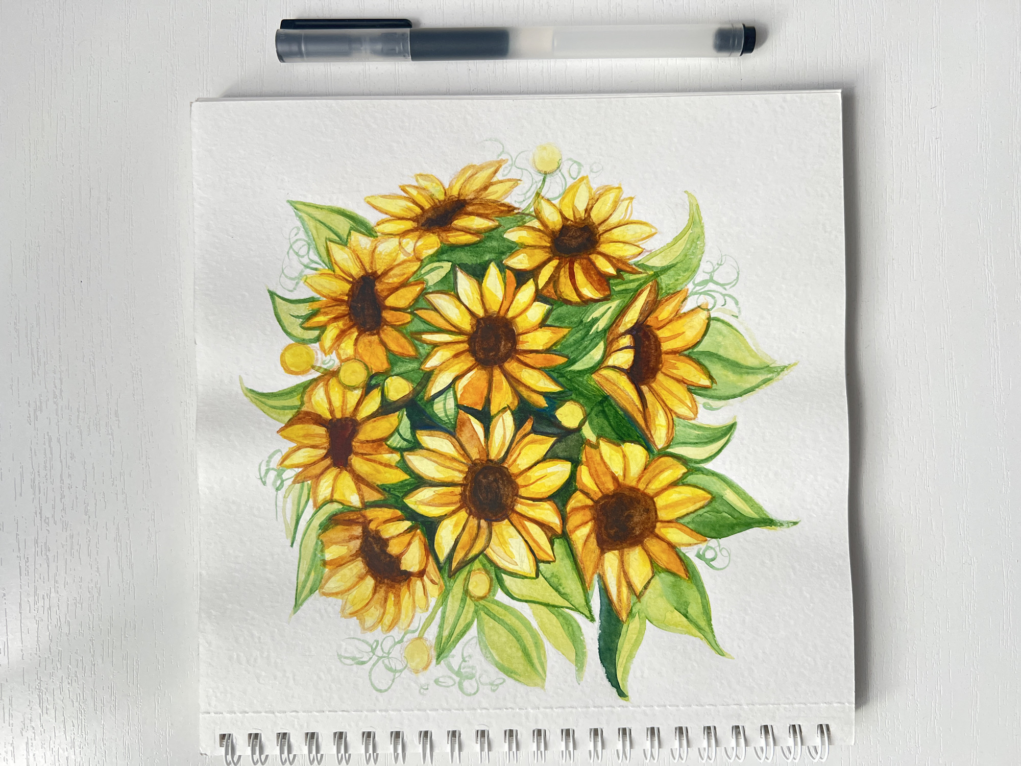 A watercolor painting of a bouquet of sunflowers on a sketchbook is shown. A pen rests above the sketchbook on a white surface. The location is impossible to determine with this image.