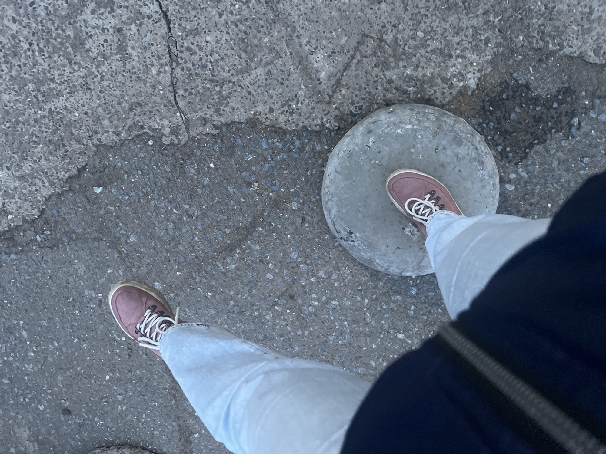 This high-angle, top-down photograph captures a person's lower body, specifically their legs and feet, on an outdoor concrete surface. The individual wears light-wash blue jeans and light pink or mauve sneakers with white laces. One foot is positioned on top of a circular, flat, grey concrete disc or stone, while the other foot rests on the surrounding ground.
The ground consists of varied concrete pavement, exhibiting different textures ranging from coarse with visible aggregate to smoother patches. Several cracks are visible in the pavement, with one prominent crack running diagonally across the upper left. Darker, potentially damp areas are also present on the concrete surface. A dark blue or black garment, possibly a jacket or bag, is visible on the right side of the frame, partially obscuring the right leg.
The lighting suggests daytime, possibly under an overcast sky or during a late hour, as no strong shadows are cast. The scene appears to be an urban or semi-urban outdoor setting, consistent with a street or pedestrian area in Chaikovsky, Russia. No specific activities beyond standing or pausing are clearly indicated, and no text is visible within the image.