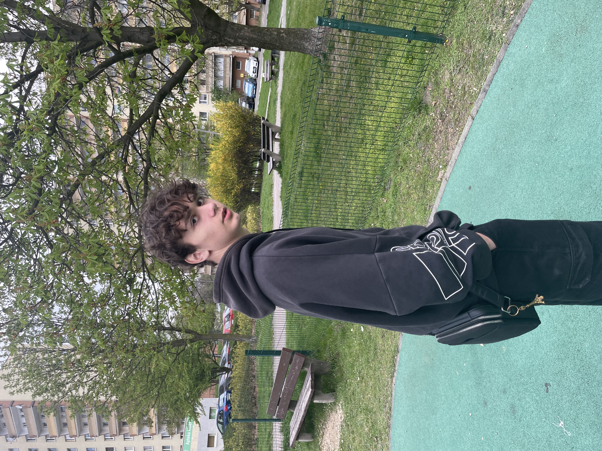 A young person with curly hair, wearing a black hoodie and dark pants, stands on a green surface in a park setting. The individual is angled towards the left of the frame and looking towards something out of view. The person has a small black bag hanging from their waist. The park has a metal fence and green grass. Behind the fence, there is a park bench, a small yellow shrub, and another bench farther in the background. Buildings are visible behind the trees, and cars are parked nearby. The location appears to be an urban park area.