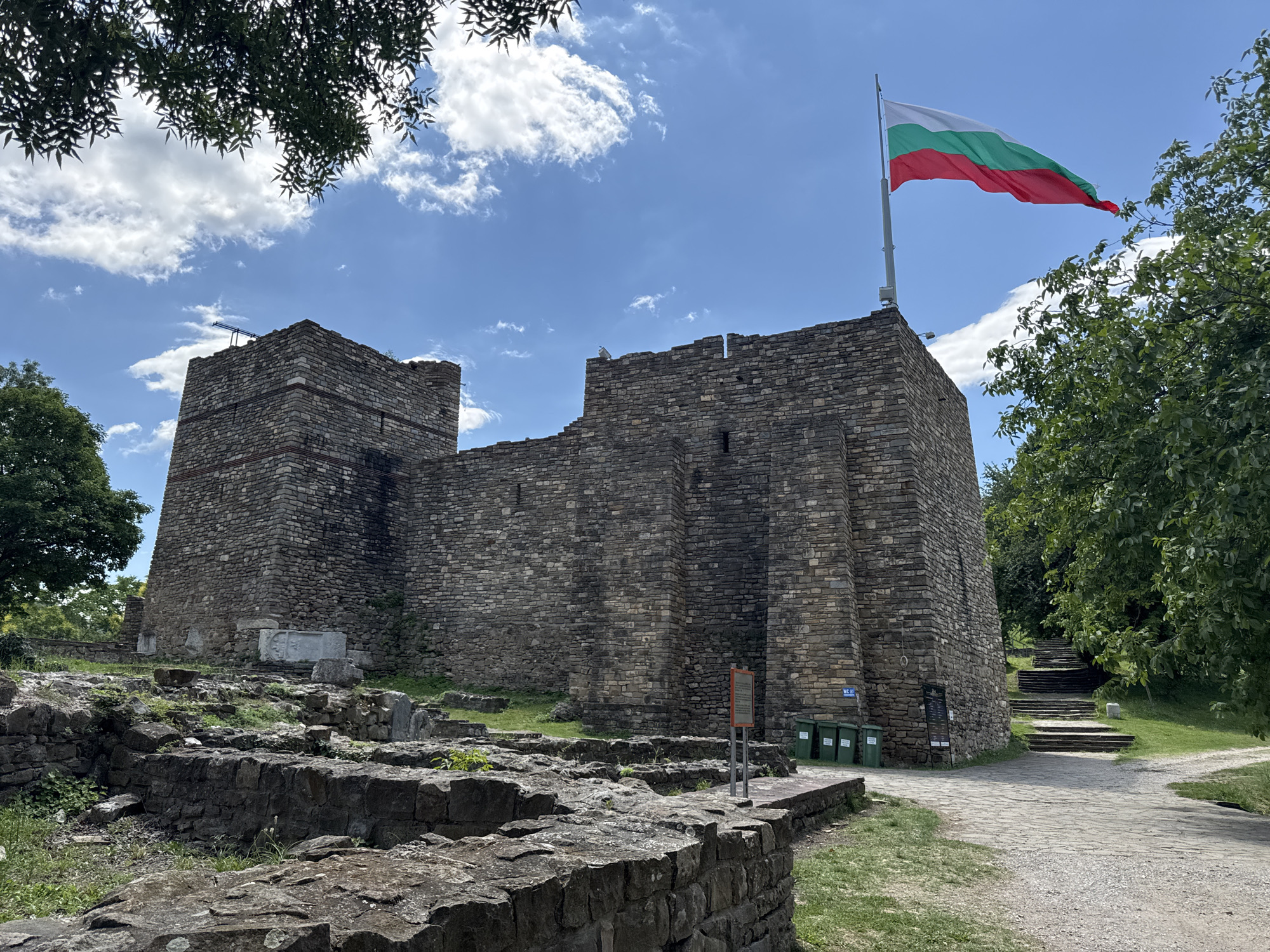 The image shows the medieval Tsarevets fortress in Veliko Tarnovo, Bulgaria. The fortress is constructed of stone and features several towers and walls. A Bulgarian flag is prominently displayed on a flagpole to the right of the fortress. The sky is blue with scattered white clouds. Trees and green vegetation surround the fortress, and a stone pathway leads up to the fortress entrance. In the foreground, there are ruins of older structures, covered in grass and moss. Several green trash bins are located near the entrance. The overall scene depicts a historic landmark on a sunny day.