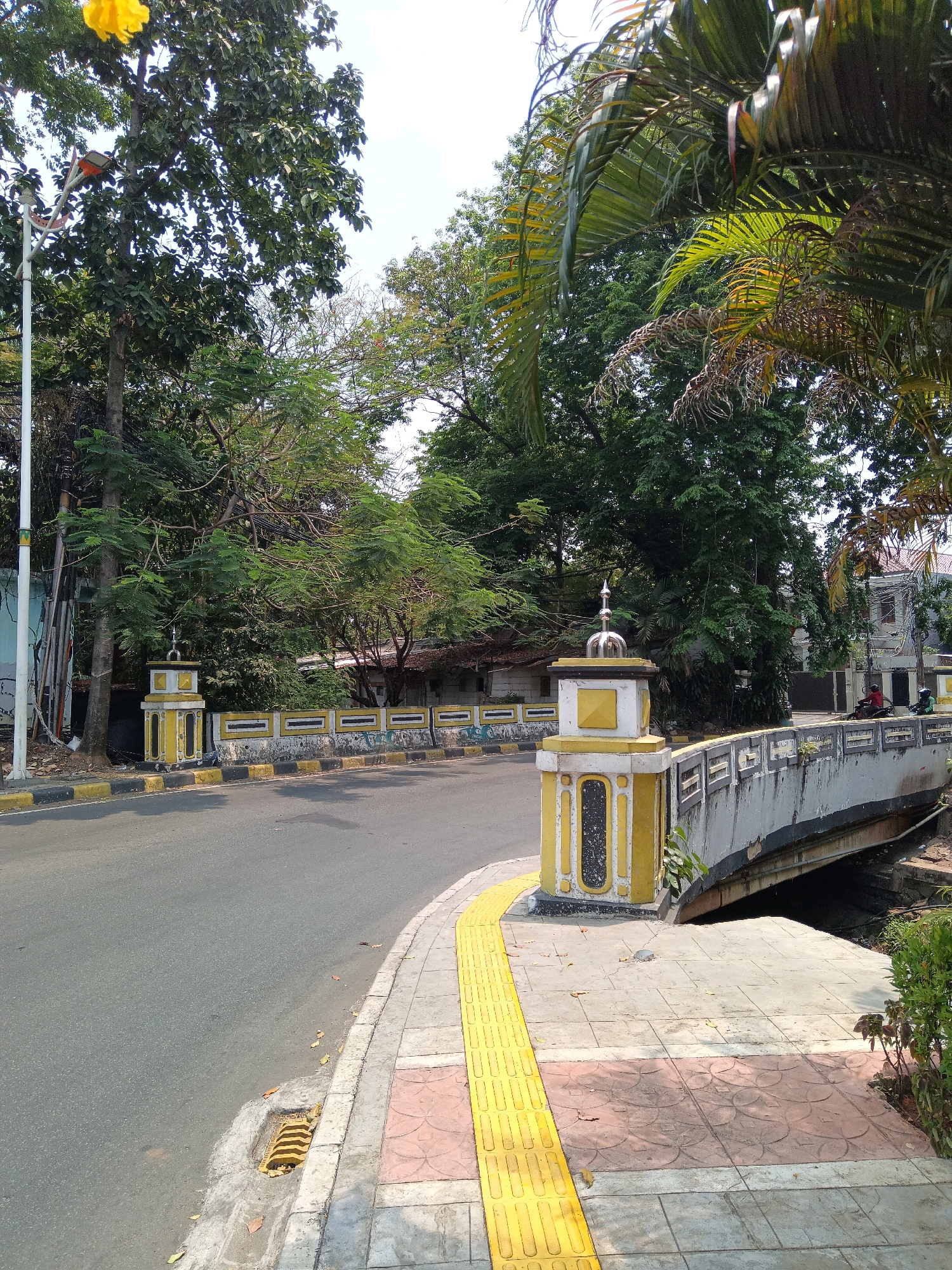 The image depicts a street scene in Central Jakarta, Indonesia. A paved road curves to the left, bordered by a sidewalk with tactile paving. To the right, a concrete bridge arches over a body of water, with a decorative yellow and white railing. The bridge and sidewalk feature ornate yellow pillars. Dense foliage from large trees lines the street and the bridge. In the background, buildings with red tile roofs are visible, and two individuals on motorcycles are present on the road. The sky is clear and bright.