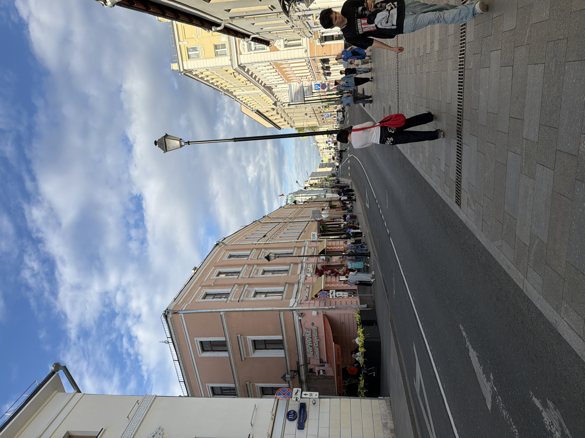 This image shows a street scene in Moscow, Russia. It is taken from street level and shows a road lined with buildings. The buildings are a mix of styles and colors, with some being yellow and others being pink. There are people walking along the sidewalk. Some of them are looking directly at the camera. There is also a lamppost in the middle of the road. The sky is blue with some clouds. Street signs in Russian are posted on the buildings. One building has Karaoke on it. The buildings show signs for Мегобарі. The street address is 11/4.