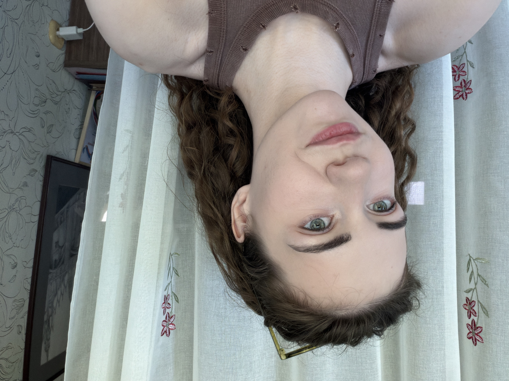 A young woman with curly brown hair and green eyes is captured in a selfie. She is wearing a brown tank top. She has light skin and is looking slightly off to the side with a subtle smile. Her hair is pulled back with a golden hair clip. Behind her, white curtains with embroidered red flowers are visible, along with a framed picture and wallpaper. The scene appears to be indoors, possibly in a home environment. There is no indication of the specific location within Zavodoukovsk, Russia, or any specific actions or interactions occurring in the image.