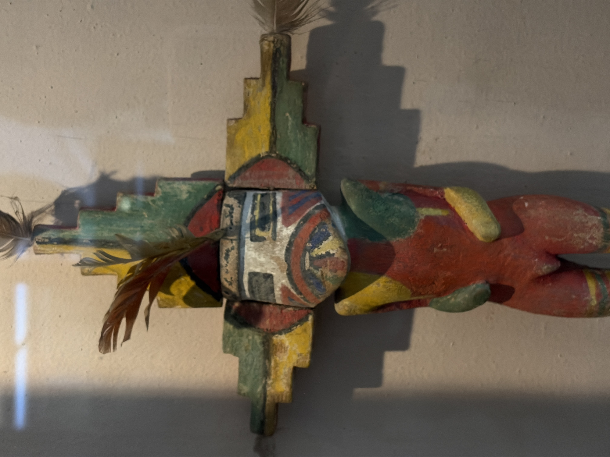 The image shows a colorful, possibly handcrafted, wooden artifact mounted on a plain wall. The artifact is roughly cross-shaped, with the central element featuring a shield-like design. Feathers are attached to the left and top of the artifact. Colors include red, yellow, green, and white, giving it a slightly aged or weathered appearance. The wall is neutral in tone, and the lighting suggests an indoor setting. There is no indication of Saint Petersburg, Russia in the image.