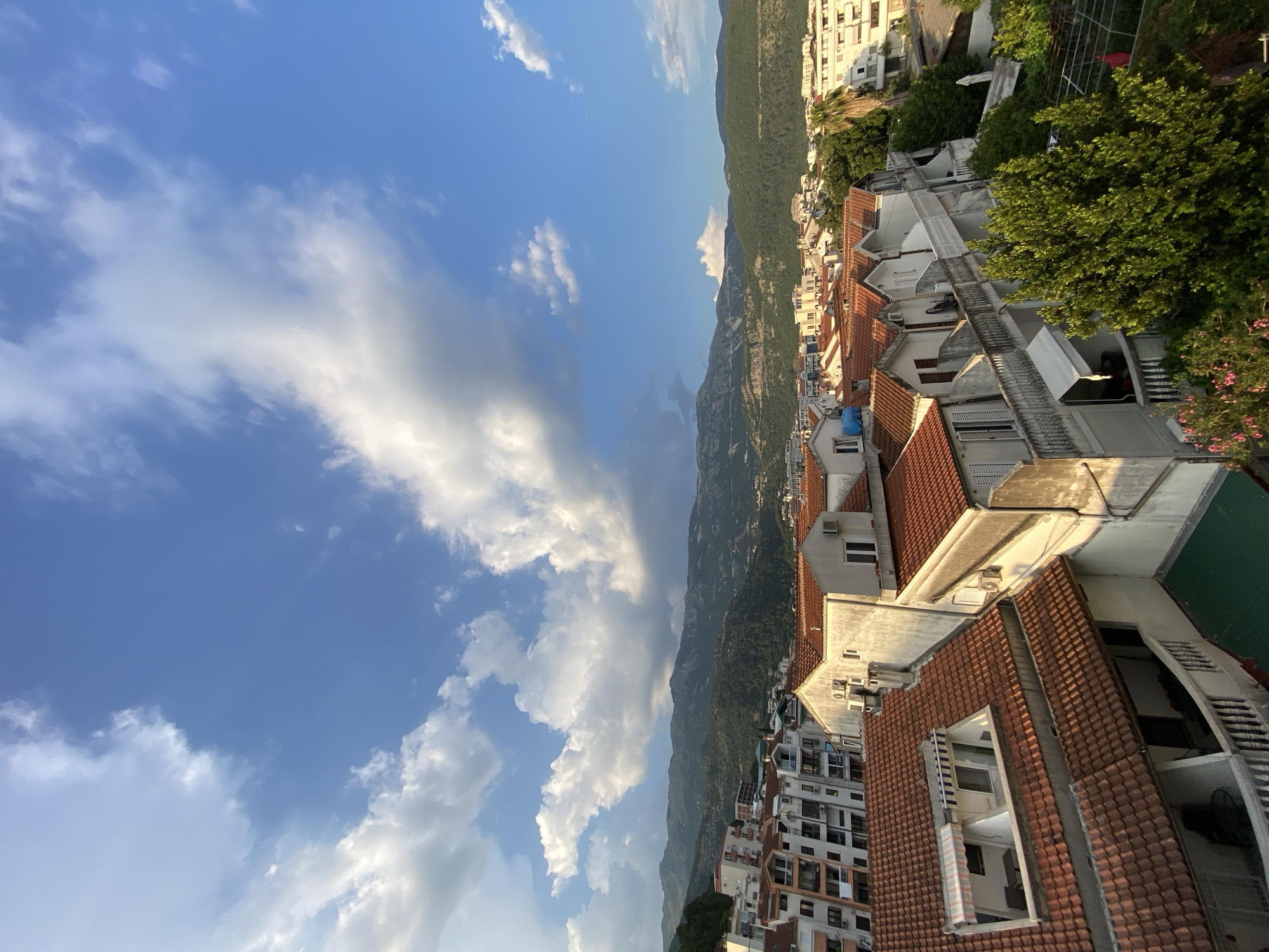 An elevated view in Budva, Montenegro depicts a partly cloudy sky covering half the scene. Below, a mountain range stretches horizontally across the middle ground. The foreground features dense urban development, with numerous buildings displaying orange tile roofs. Vegetation is interspersed amongst the buildings, more concentrated on the right side of the frame. The photograph is shot with a significant degree of rotation.