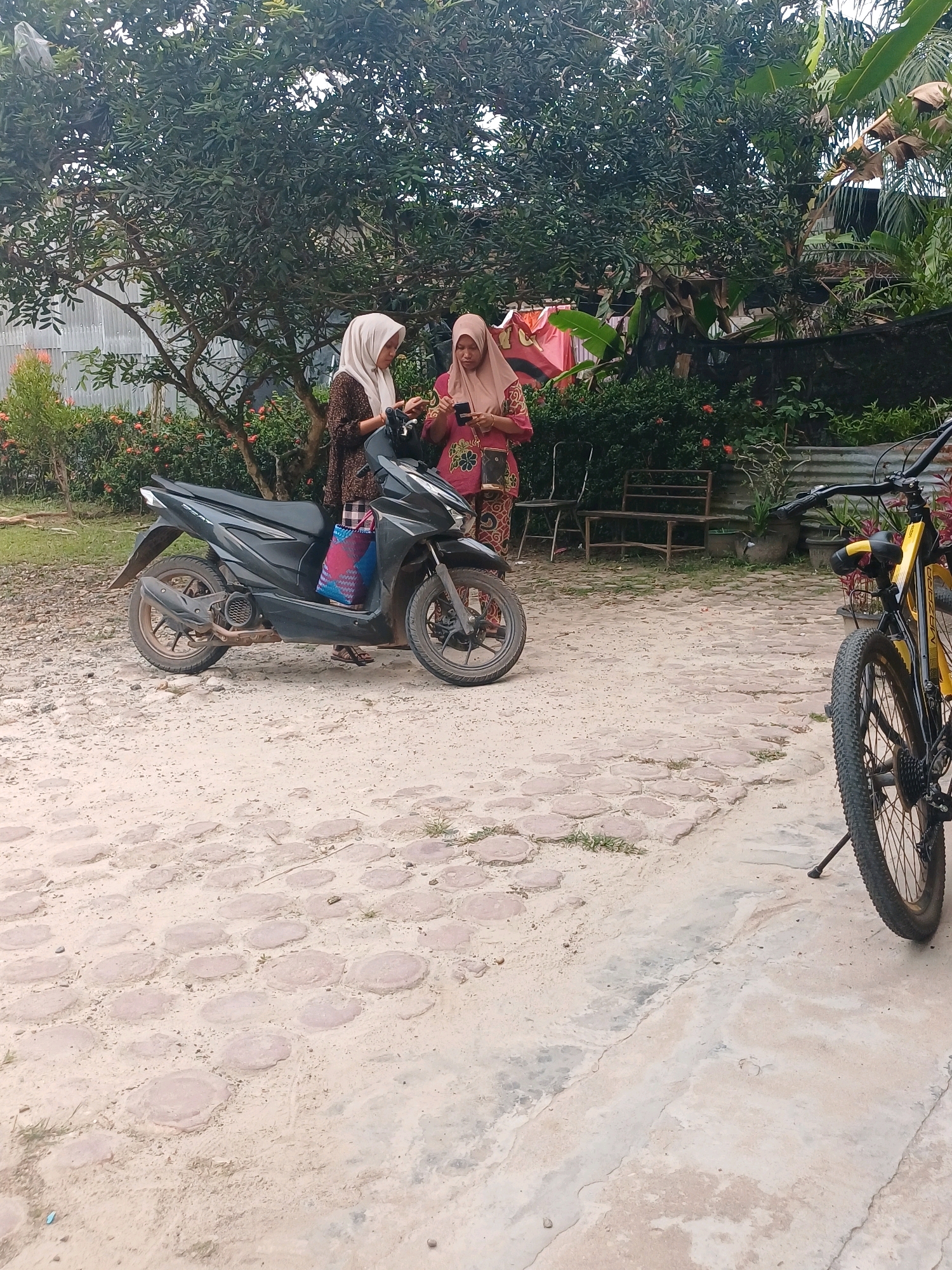 In Dumai City, Indonesia, two young women wearing hijabs are standing next to a black motorcycle. One woman, wearing a light brown hijab and a patterned brown and white dress, is holding a smartphone. The other woman, with a pinkish hijab and a patterned pink and white dress, is also looking at her smartphone. A blue and pink patterned bag is placed on the seat of the motorcycle. To the right of the motorcycle, a yellow and black bicycle is parked, with its kickstand down. The scene is set on a dirt and stone-paved ground, with lush green trees and foliage in the background, suggesting a rural or semi-rural residential area. A corrugated metal fence is visible behind the trees.