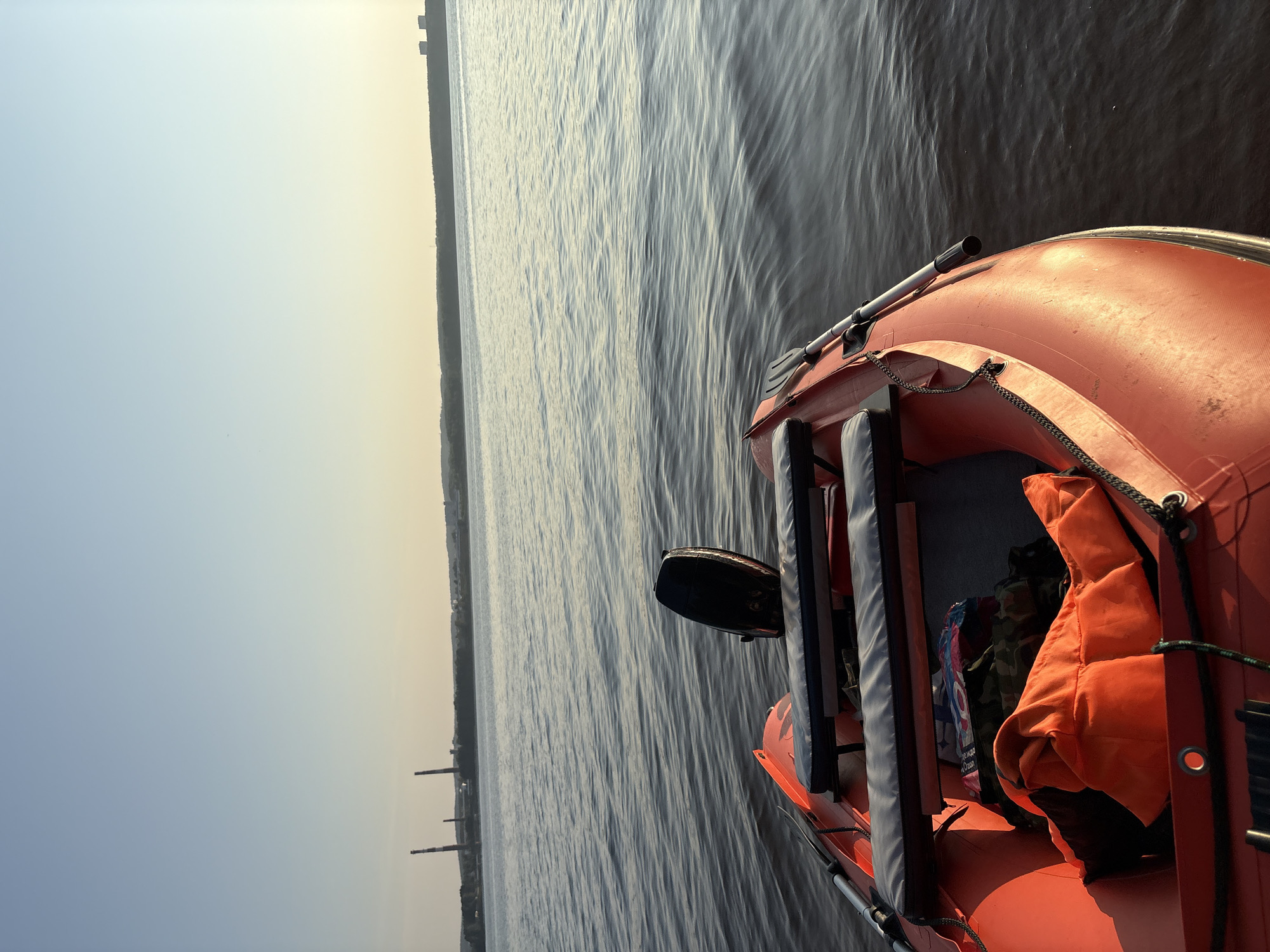 An overhead perspective shows an orange inflatable boat on the Kama River near the city of Chaykovsky, Russia. The boat is equipped with a black outboard motor, two padded seats, and a life jacket in bright orange. The river water is calm, with small ripples. In the distance, the hazy outlines of industrial smokestacks and buildings can be seen along the shoreline. The sky is clear and pale blue, transitioning to a warm yellow hue near the horizon, indicating either sunrise or sunset.