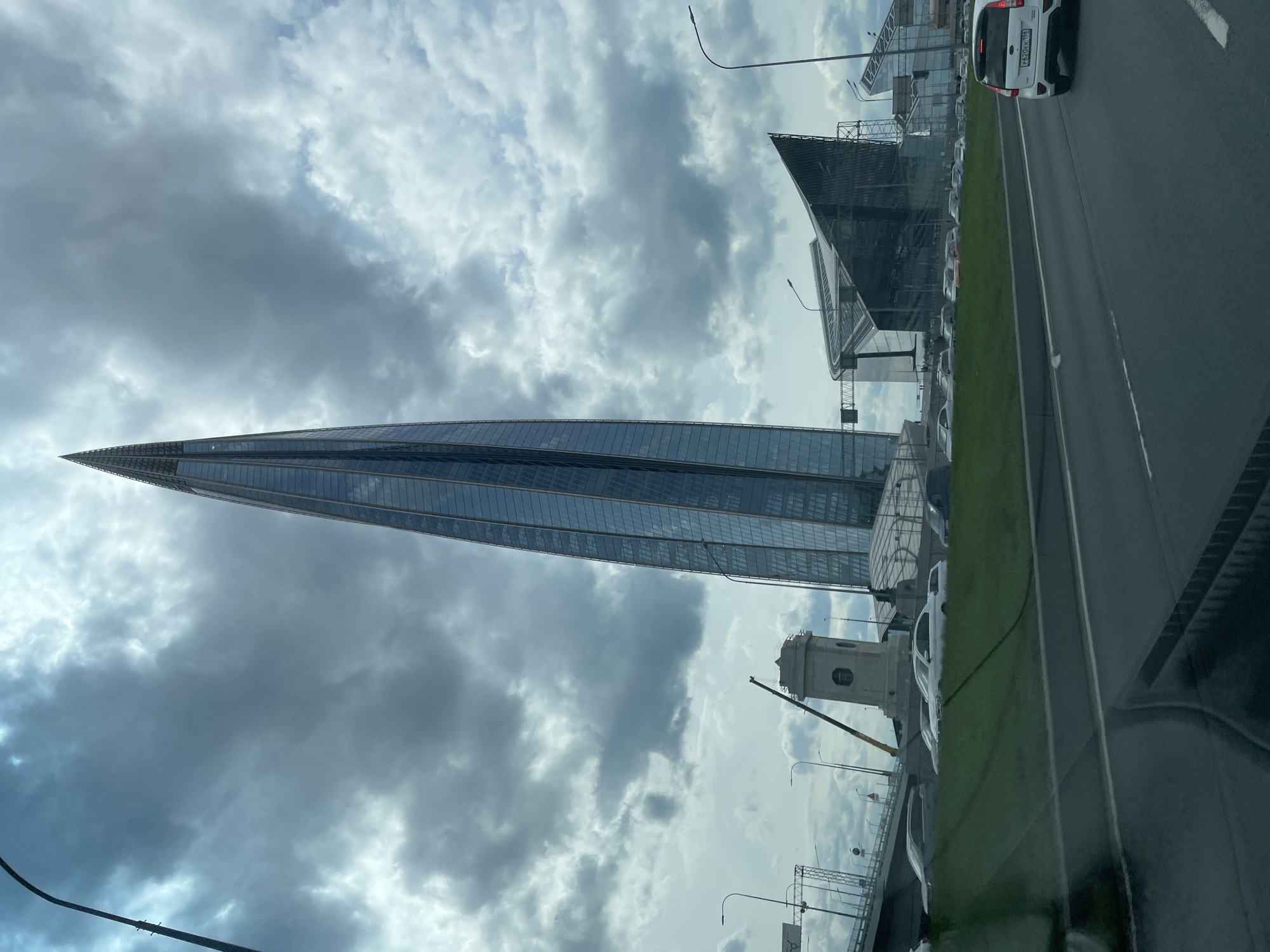The image shows a view of the Lakhta Center in Saint Petersburg, Russia, as seen from a vehicle. The Lakhta Center is a modern skyscraper with a pointed, spire-like design. The sky is overcast with grey clouds. In the foreground, there is a road with vehicles. Along the roadside, there's some greenery separating the road from other buildings, including the under construction new railway station. There is a white car visible on the road with a partial view of its license plate. The image seems to have been taken from inside the vehicle.