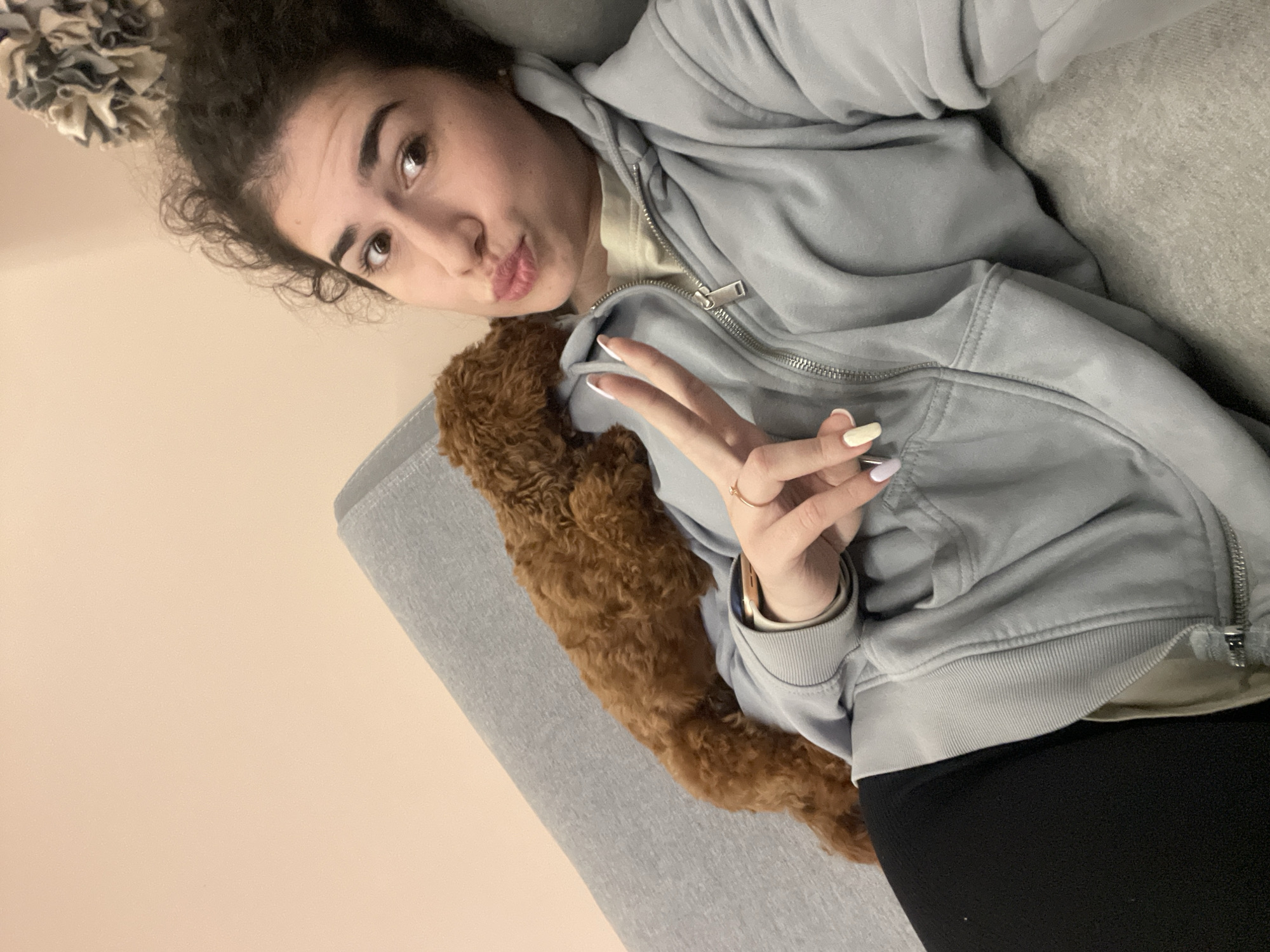 A woman is lying on a gray sofa, posing for a selfie with a brown poodle lying on her chest. She is wearing a light blue zip-up hoodie over a cream-colored top and black leggings. Her nails are painted in pastel colors, and she is making a peace sign with her right hand. The background consists of a beige wall. The image does not provide any information related to the city of Sofia, Bulgaria.