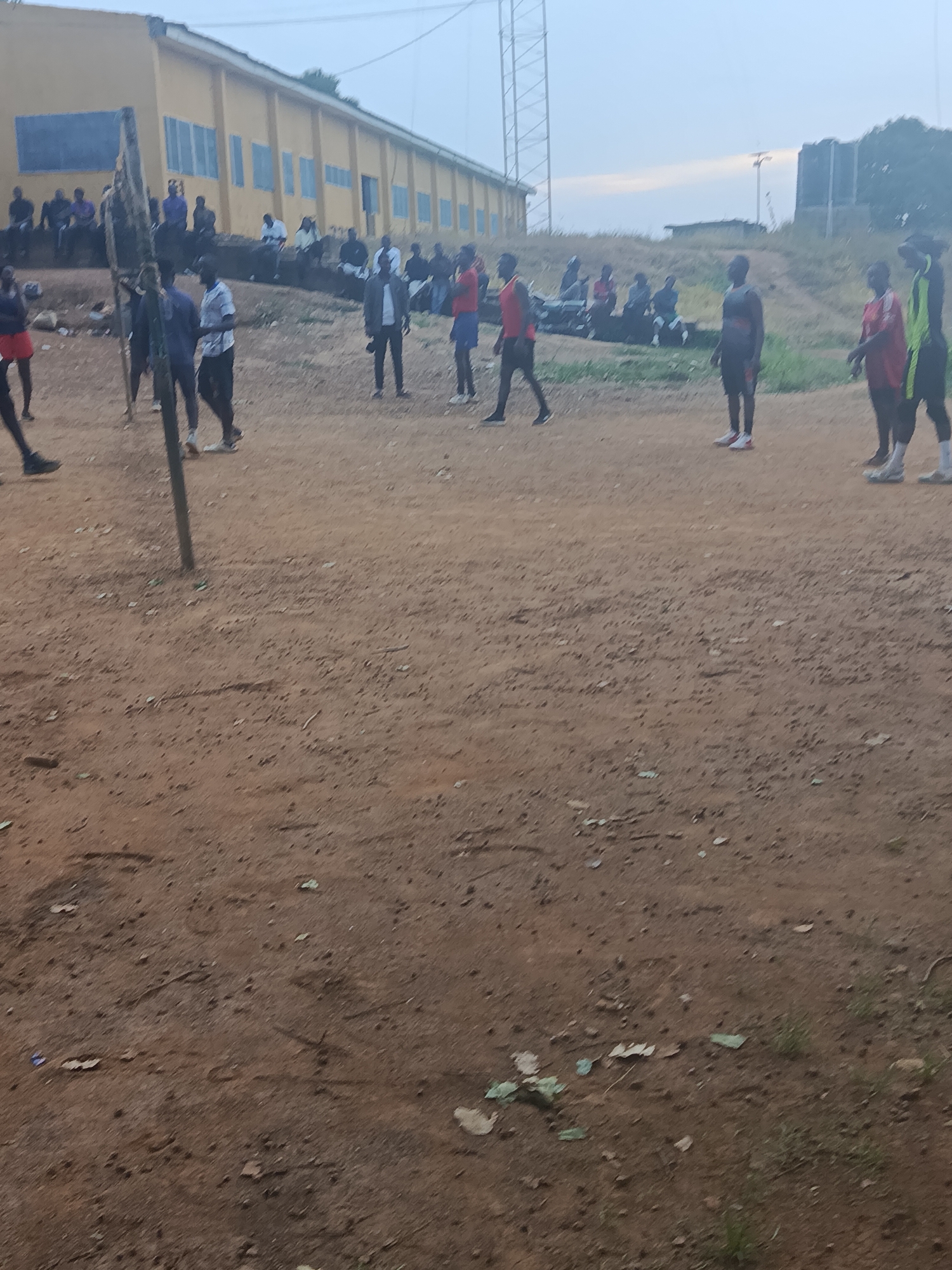 The image depicts an outdoor scene, likely a sports field, in Rafin Par, Nigeria. In the background, a long, yellow, two-story building with blue-framed windows is visible. Several individuals are gathered on and around the building's steps and in front of it, appearing to be spectators or participants. The foreground is dominated by a dirt or sand ground, suggesting a volleyball or similar court. A vertical pole, possibly part of a volleyball net, is positioned prominently in the left-center of the frame. Several young men, dressed in athletic attire such as shorts and jerseys, are scattered across the field. Some are standing, while others appear to be in motion, possibly playing a game. The lighting suggests it is either early morning or late evening, with a hazy sky. The overall environment is rustic, with sparse vegetation and debris visible on the ground.