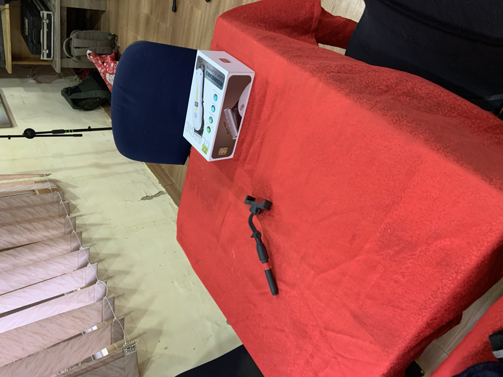 The image depicts an indoor scene, possibly a room or a storage area. A table covered with a vibrant red tablecloth is the central focus. On the table, there are two objects: a black and red handheld device, likely a selfie stick or a similar accessory, and a white rectangular box which appears to be packaging for a hair clipper. The box is labeled "CLASSIC" and "RECHARGEABLE HAIR CLIPPER."
The background reveals elements that suggest a functional space. To the left, vertical blinds with a patterned design cover a window. The wall behind the blinds is a pale yellow and shows signs of wear and tear. Further back, a dark blue chair is partially visible. To the right of the chair, a tall black microphone stand is positioned. Behind the microphone stand, a dark green backpack rests against the wall. Another olive-colored backpack is situated near a wooden shelf unit in the far right of the image, on which a black case is placed. A red and white patterned object, possibly a gift wrap or fabric, is also visible near the backpacks. The floor appears to be wooden. The overall impression is one of a somewhat cluttered but organized space. The location of Gura Top, Nigeria, is provided contextually, but the image itself does not contain any specific visual cues to confirm this geographical placement.
