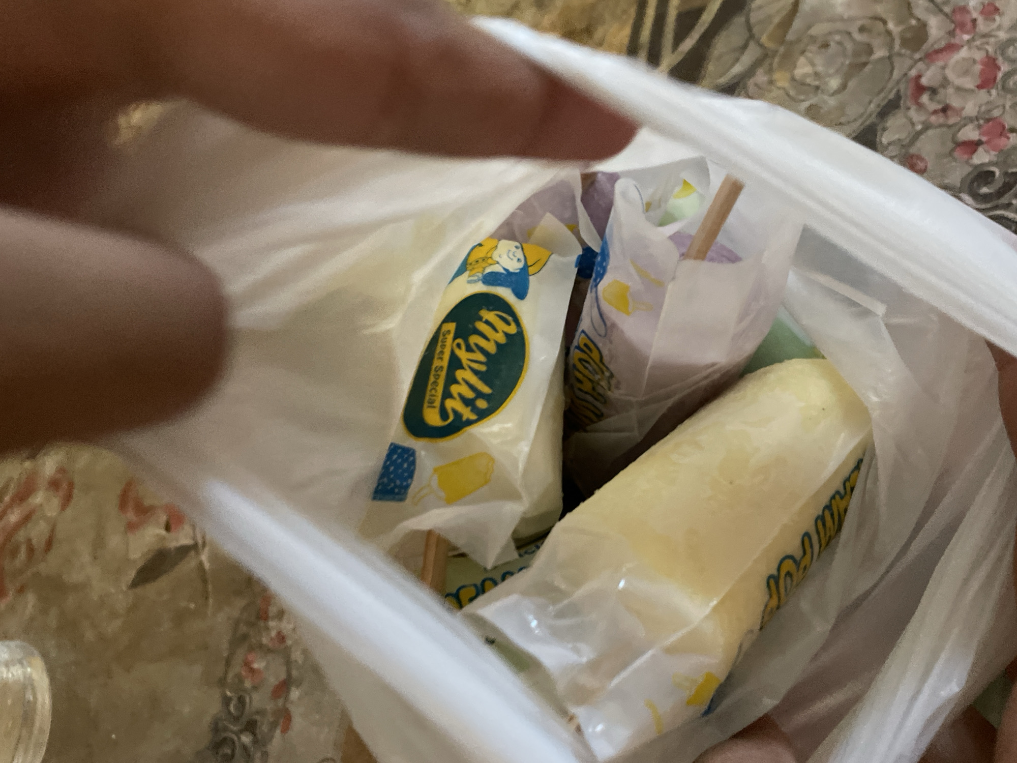 A white translucent plastic bag is held open by human hands, revealing at least three individually wrapped frozen treats on wooden sticks. The most prominent treat is a light yellow/cream-colored ice pop, enclosed in clear plastic packaging labeled "MyLit Super Special" with green and yellow text, featuring a blue-capped cartoon character and a yellow beverage icon. Below this, a second treat with a partially visible purplish wrapper and a protruding wooden stick is present, alongside a potential third item wrapped in green. Human fingers are partially visible at the upper right and lower left, gripping the bag. The background consists of an intricately patterned surface, possibly a tablecloth or tiled floor, displaying abstract or floral motifs in shades of light brown, grey, and faded pink. The scene is located indoors in Davao City, Philippines.