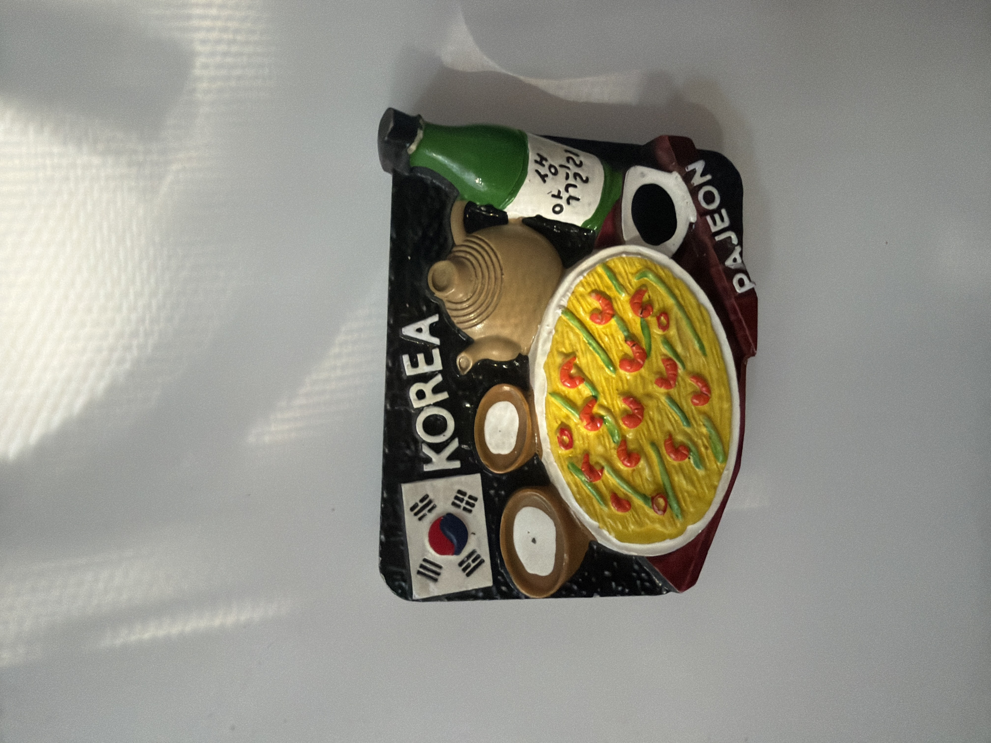 This is a close-up, overhead shot of a novelty refrigerator magnet depicting Korean culture. The magnet is rectangular with rounded corners and features a black background. In the upper left corner is the South Korean flag. To its right, the word "KOREA" is printed in white capital letters. Below the flag are two small brown bowls with white centers, possibly representing dipping sauces. The central portion of the magnet displays a round, yellow dish with red shrimp and green scallions, resembling a Korean pancake called Pajeon. To the right of the pancake is a light brown teapot with a lid. Further to the right is a green bottle with a black cap, featuring Korean text in black on a white label. Below the bottle is a small white cup with a black interior. At the bottom of the magnet, the word "PAJEON" is printed in white capital letters. The magnet is positioned on a plain white surface, possibly a refrigerator door, with light and shadow patterns cast from an unseen light source above. The location in Shymkent, Kazakhstan cannot be determined from the image.