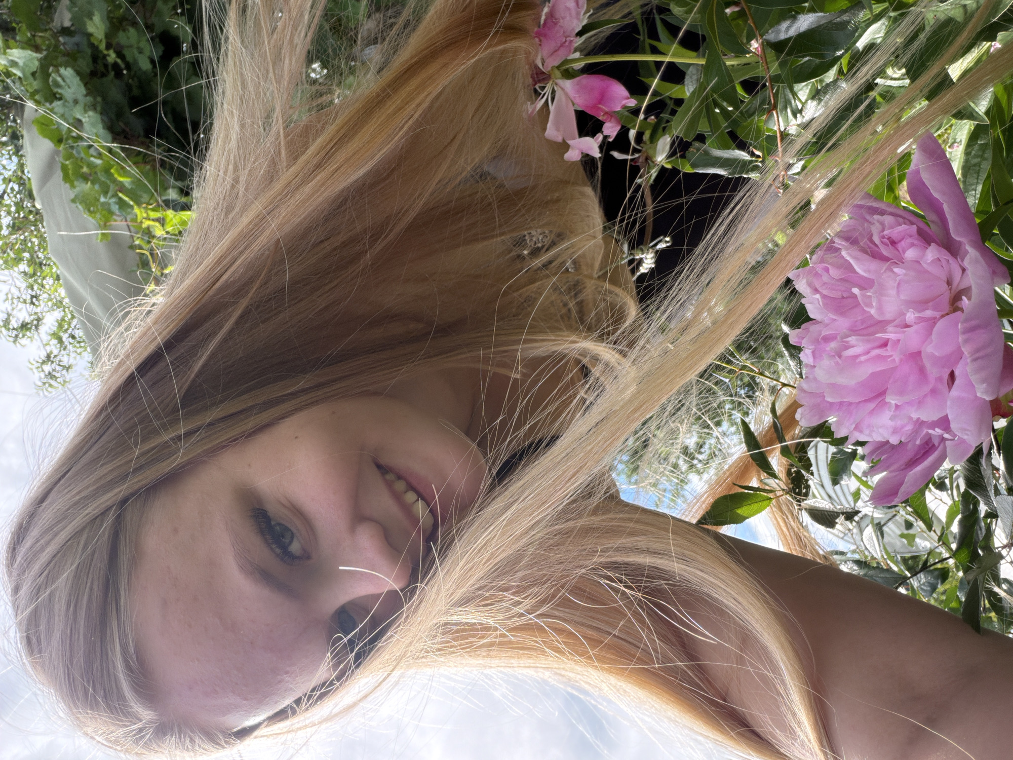 A young woman with blonde hair is captured in a selfie taken outdoors. She is smiling with her mouth open, showing her teeth, and her eyes are looking towards the camera. Her hair is long and blonde, and some strands are falling across her face. She is surrounded by greenery and pink peonies, suggesting a garden or natural setting. The lighting indicates it is daytime, likely sunny, given the brightness and the way light catches her hair. The image is taken from a low angle, looking up at the woman. The overall mood appears cheerful and natural.