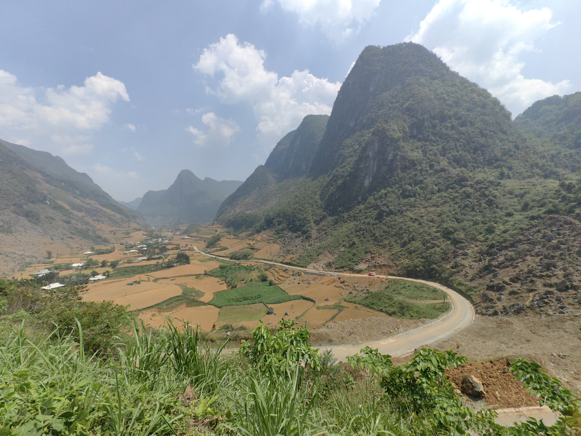The image depicts a valley landscape in Xã Xuân Trường, Vietnam. Towering, densely forested mountains frame a valley with agricultural fields and a winding road. The fields are predominantly a brown-yellow hue, likely post-harvest, interspersed with patches of green vegetation. A road cuts through the valley, curving sharply in the foreground. A few buildings are visible, scattered among the fields. In the immediate foreground, there's lush green grass and shrubs. The sky is light blue with scattered white clouds. A single vehicle, possibly a truck, is visible on the road. The overall scene suggests a rural and mountainous environment.