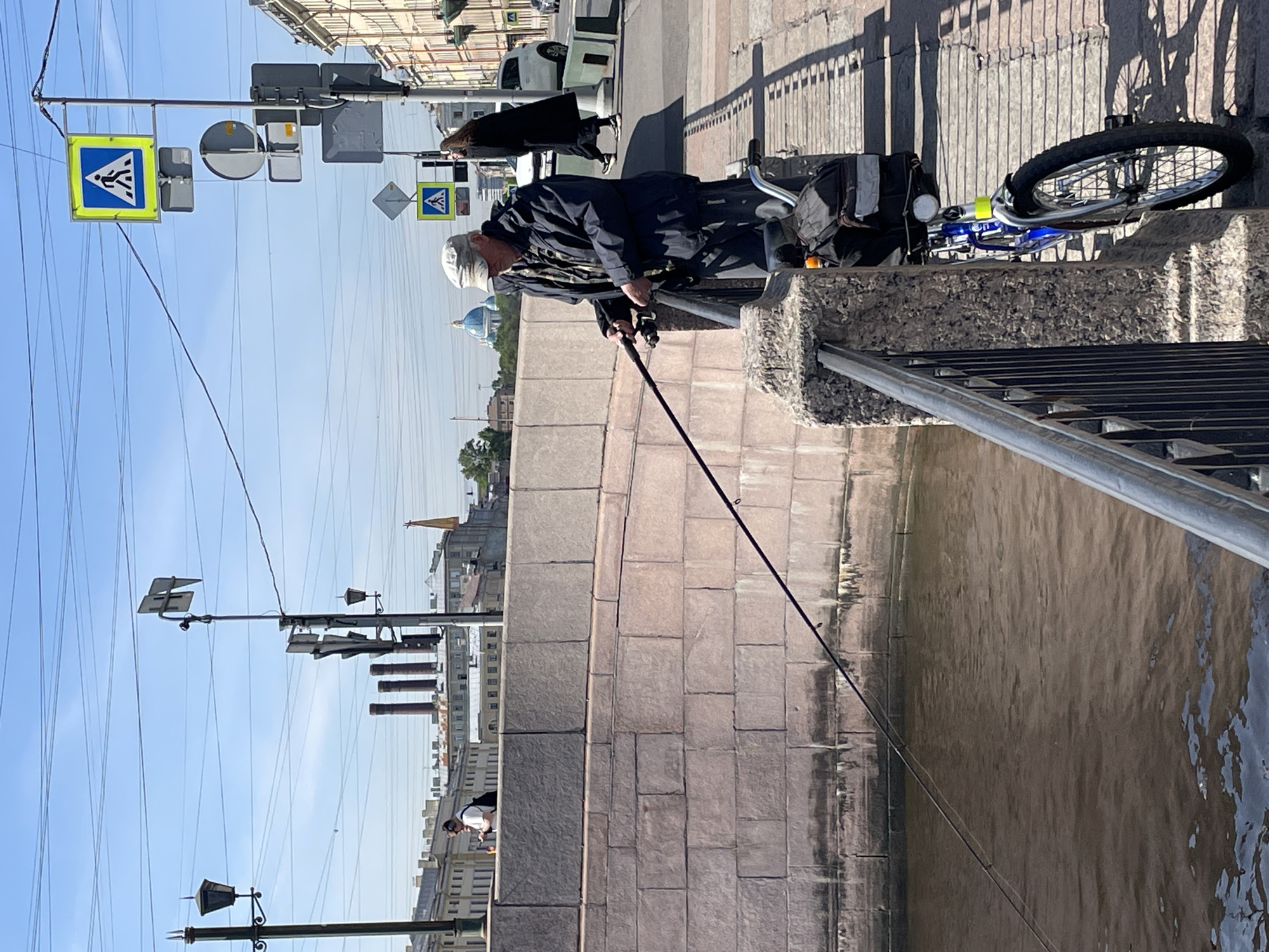 A man fishes off the side of a bridge in St. Petersburg, Russia. He is wearing a dark jacket and a white cap. A bicycle is parked next to him. The bridge is made of gray stone. A pedestrian crosswalk sign is visible in the background. Across the water, city buildings are visible. The day is sunny with a blue sky.