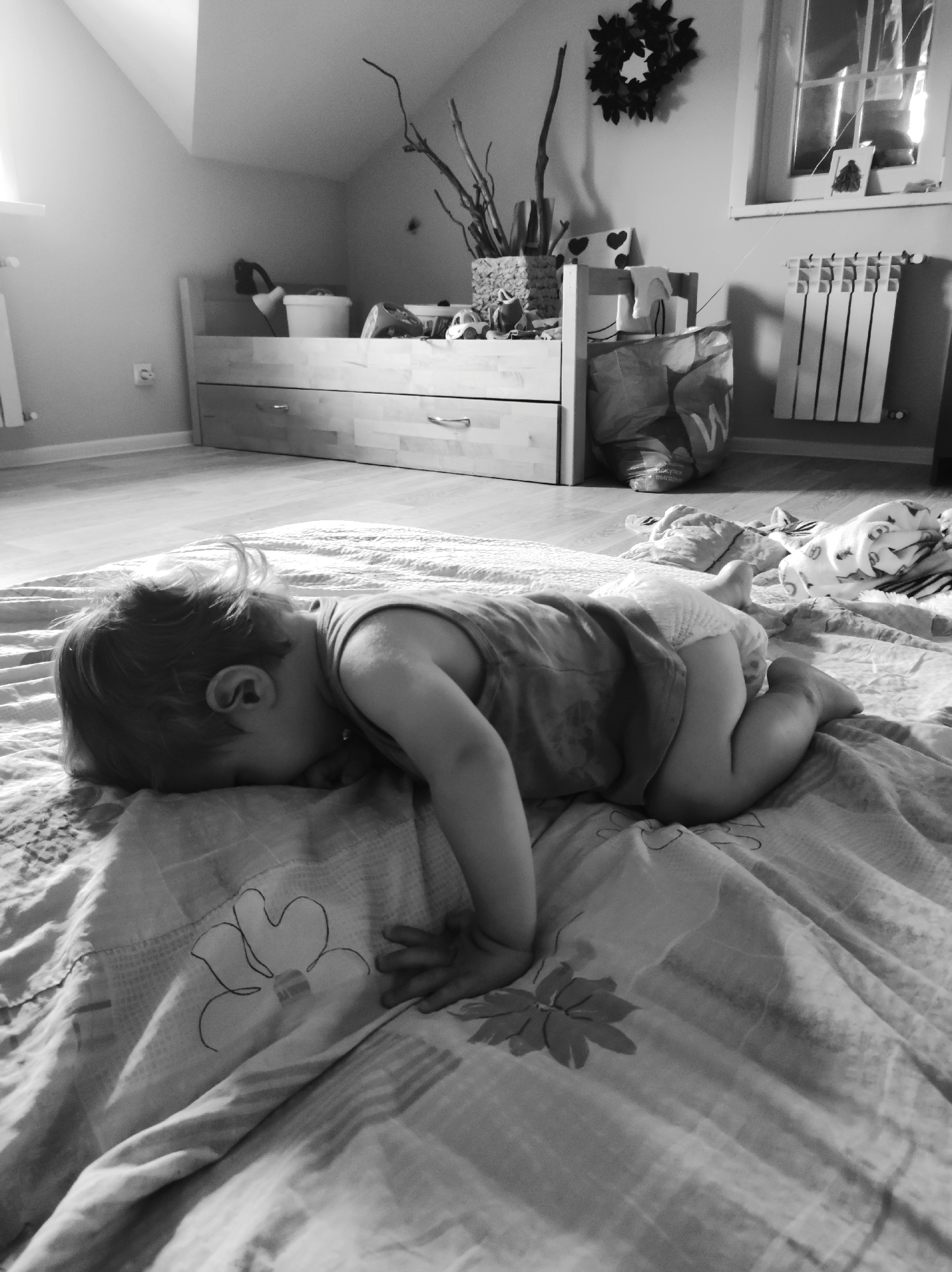 The black and white photograph captures an interior scene, likely a child's bedroom or nursery, in the city of Unknown, Unknown. A young child, appearing to be a toddler, is positioned in the foreground, lying on their stomach on a light-colored, patterned blanket or duvet. Their head is turned to the left, partially obscured by the bedding, with light hair visible. The child wears a light-colored sleeveless top and a diaper, with arms bent and legs tucked underneath them. The blanket features outlines of flowers.
In the midground, a light-colored wooden furniture unit, resembling a low bed frame with two drawers, occupies the central space. The top surface of this unit serves as a shelf, displaying various items: from left to right, a dark object possibly a lamp base, a white bucket, an alarm clock with a round face, and a small toy car. Behind these items, a vase containing several dried tree branches is visible. A patterned pillow with heart shapes is also present on the shelf.
The room features light-colored walls, possibly white or light grey. On the left side of the frame, a window lets in bright light, and a white, vertical radiator is mounted below it. A white electrical outlet is positioned on the wall between the radiator and the wooden furniture. The floor consists of light-colored wooden planks with a matching baseboard.
On the right side of the background, another window with multiple panes is visible, reflecting bright light. Below this window, a smaller white radiator is installed. A dark, circular wreath or decorative object hangs on the wall above the wooden furniture unit. A large, light-colored fabric bag with a dark "W" logo and other text (possibly "скидки" or "выгодные покупки" in Cyrillic) rests on the floor next to the wooden furniture unit. Behind the child's feet, a crumpled blanket or towel and other soft items are visible on the floor. The ceiling on the left side of the room appears sloped, suggesting an attic space or room under an eaves.