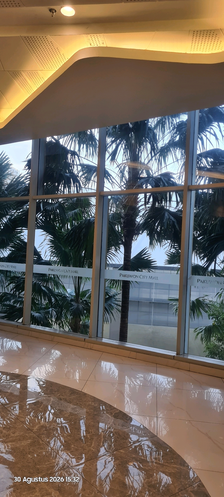 The image shows an interior view of a building, likely a shopping mall or hotel, with large windows overlooking palm trees and a building facade. The floor is polished and reflects the scene outside. The text "PAKUWON CITY MALL" is visible on the windows. A timestamp "30 Agustus 2026 15:32" is in the bottom left corner, indicating the date and time the photo was taken, which is August 30, 2026, at 3:32 PM. The ceiling has integrated lighting and what appears to be a fire sprinkler head. The scene suggests an indoor environment in a tropical or subtropical location, given the presence of palm trees. Pakuwon City Mall is a known shopping mall located in Surabaya, Indonesia.