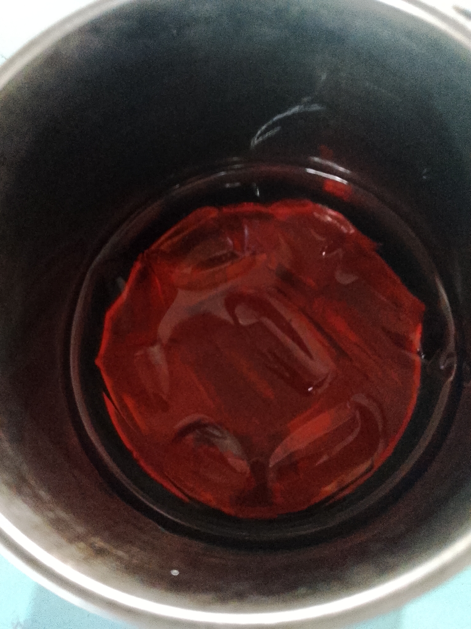 A top-down view shows a stainless steel pot containing a mass of red gelatin submerged in a clear liquid, likely water. The gelatin has an irregular, wrinkled surface. The inside of the pot is dark gray to black. The outer rim of the pot is visible, along with a pale blue or green surface beneath it. The location is unidentifiable as Lhokseumawe, Indonesia from this image alone.