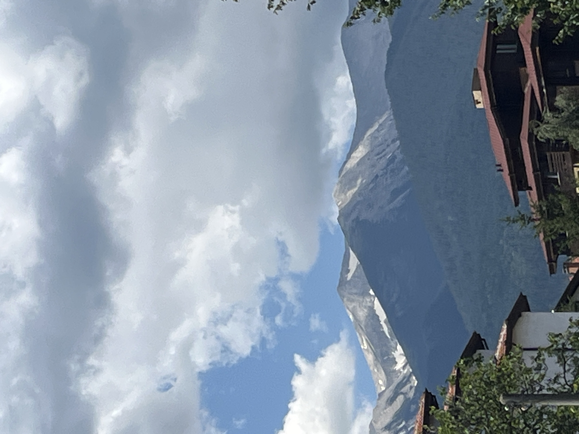 The image depicts a scenic view of the Pirin Mountains, likely taken from the city of Bansko, Bulgaria. The foreground shows the roofs of traditional Bulgarian houses, characterized by red-tiled roofs and white walls, with lush green trees partially obscuring the structures. In the mid-ground, a dense forest covers the lower slopes of the mountains. The upper slopes of the Pirin Mountains are visible, with patches of snow remaining on the jagged, rocky peaks, suggesting it might be late spring or early summer. The sky is a mix of blue sky with white fluffy clouds and darker, overcast clouds, indicating dynamic weather conditions. There are no people or animals visible in the image. The overall scene conveys a sense of natural beauty and the peaceful coexistence of human settlements with a grand mountain landscape.