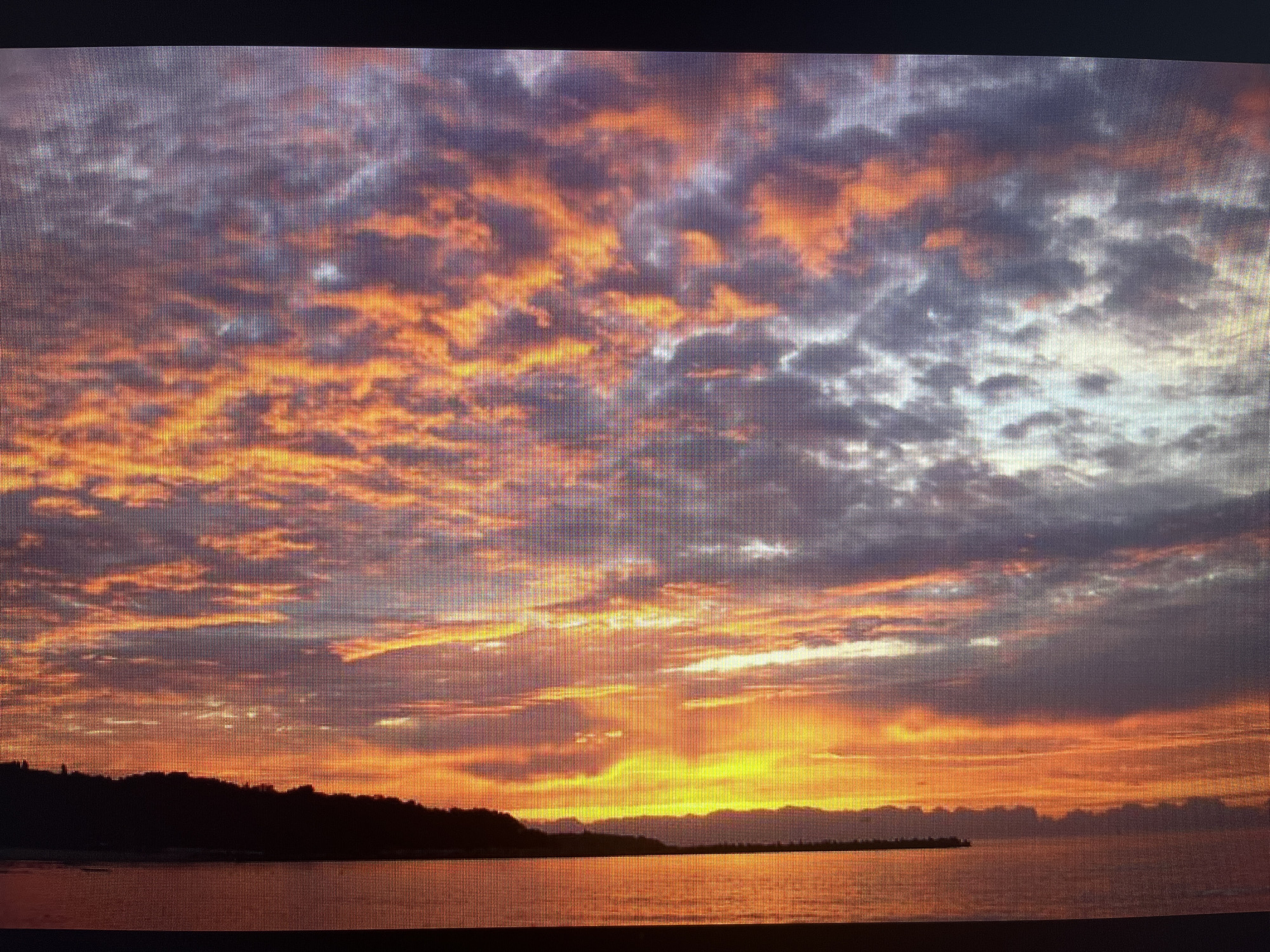 A vibrant sunset is depicted over a calm body of water, likely a sea or large lake, with a distant landmass silhouetted against the horizon. The sky is the dominant feature, filled with dramatic, scattered clouds illuminated in an array of warm colors.
The upper and central sky display rich tones of fiery orange, deep red, and bright yellow, particularly where the sun's rays break through the clouds near the center-right. These warm hues blend upwards and to the right into cooler purples, grays, and softer whites. The clouds themselves are diverse, ranging from thin, wispy streaks to denser, puffier formations, all catching the intense light of the setting sun.
Below the colorful sky, a dark, undulating silhouette of land stretches horizontally across the middle of the frame. On the left, it appears as a tree-covered coastline, while towards the right, it becomes a lower, less defined landmass or perhaps a distant breakwater extending into the water.
The water's surface acts as a mirror, reflecting the brilliant oranges, yellows, and reds of the sky, creating shimmering paths of light that fade into darker, reddish-brown tones further from the sun's reflection. The water appears tranquil, with minimal discernible waves.
The time of day is clearly sunset, characterized by the low sun and the intense, warm light. The overall atmosphere is serene and awe-inspiring. The image itself exhibits a distinct grid-like pixelation across its entirety, suggesting it is a photograph taken of a digital screen or display. There is no visible text in the image. Given the location context, this is a sunset over the Black Sea near Varna, Bulgaria.