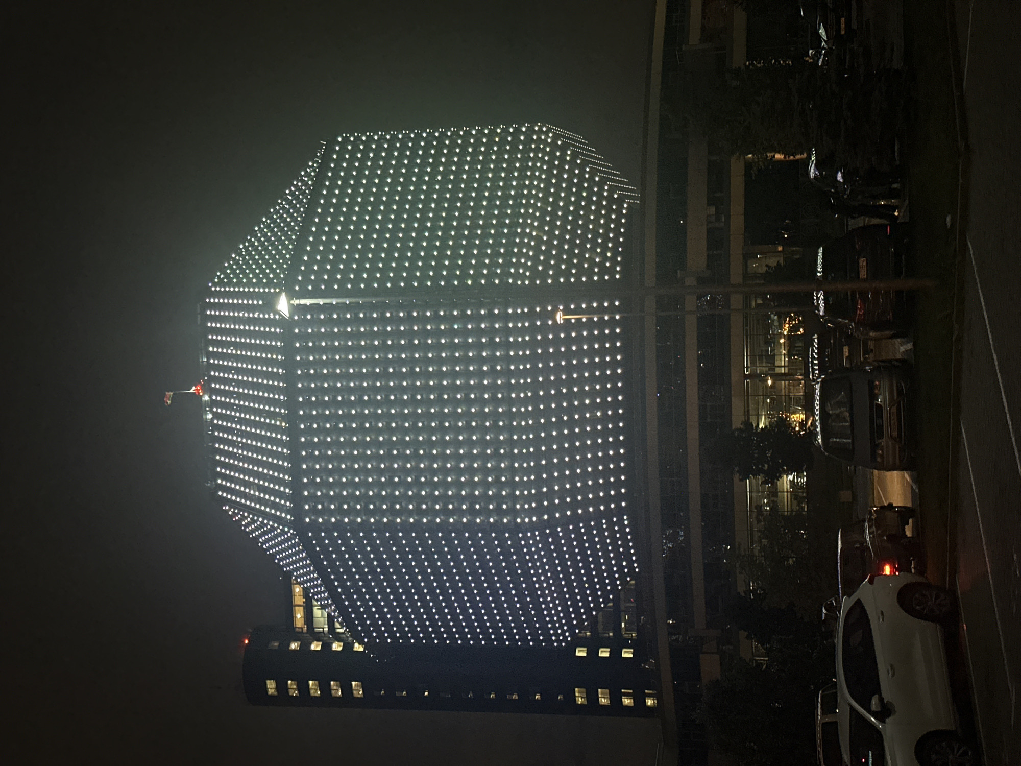 An external view of a large, multi-story building illuminated by numerous white lights, creating a patterned facade, is captured at night. The building appears to have a hexagonal or octagonal shape and is topped with a small flag. Adjacent to it, on the left, is a taller, cylindrical structure with illuminated windows. In the foreground, a parking lot is visible with several cars parked. The scene is dark, with ambient light coming from the building and streetlights, casting shadows and creating a slightly hazy atmosphere. The image is taken from a low angle, looking up at the main structure.