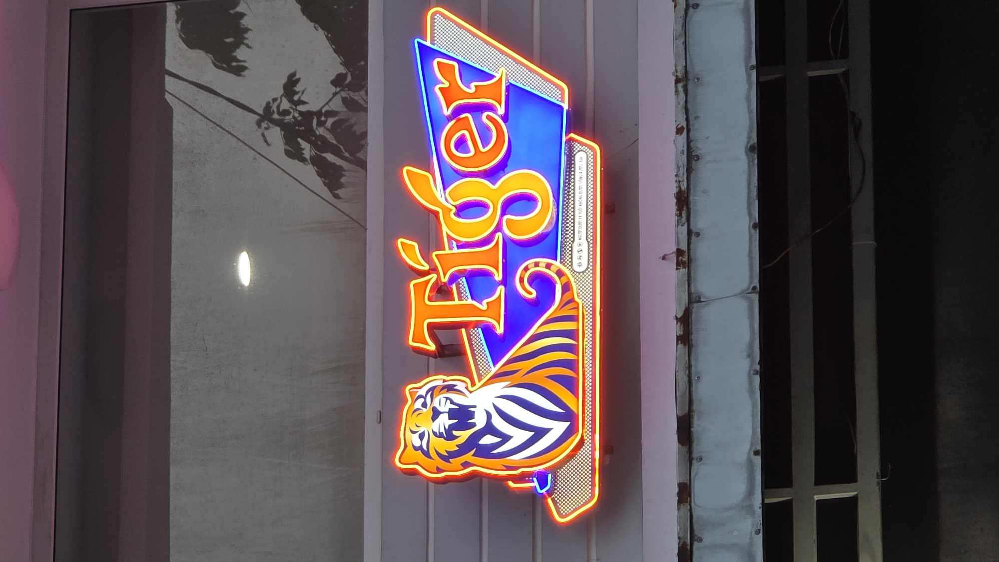 A vertical, rectangular, neon-lit sign for “Tiger” hangs outside in Nha Trang, Vietnam. The sign is affixed to a pale grey wall. The word “Tiger” is spelled out vertically in orange neon, above an image of a crouching tiger with blue and orange stripes. The sign itself is made up of a blue trapezoid outlined in orange neon. The sign is illuminated and partially obscures the gray wall and adjacent window with a tree branch reflection. To the right, part of a dark gray metal frame is visible.