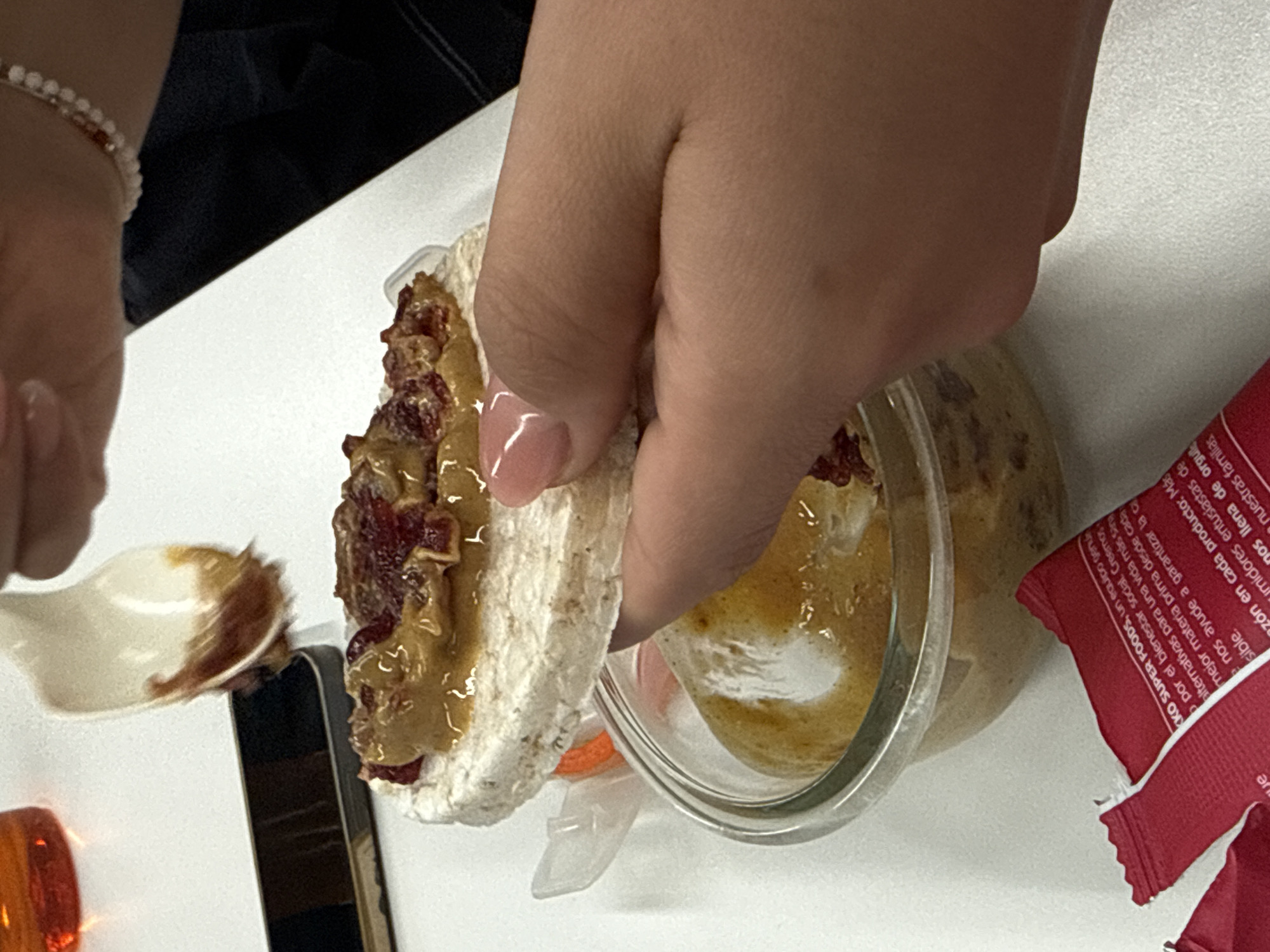 Here is a description of the image:
A close-up shot depicts a person's hand holding a rice cake topped with peanut butter and what appears to be cranberry sauce. The rice cake is slightly cracked. Next to it, a jar with a brownish liquid sits on a white surface. A small spoon with a brown-colored smear is also visible. A red packet is partially visible on the right side. The lighting suggests an indoor setting. The text on the red packet is written in Spanish and mentions "KKO SUPER FOODS" and a team dedicated to social well-being. This suggests a meal preparation or consumption context, possibly in a domestic or office setting. The city of Mexico City, Mexico is not mentioned or shown in the image.
