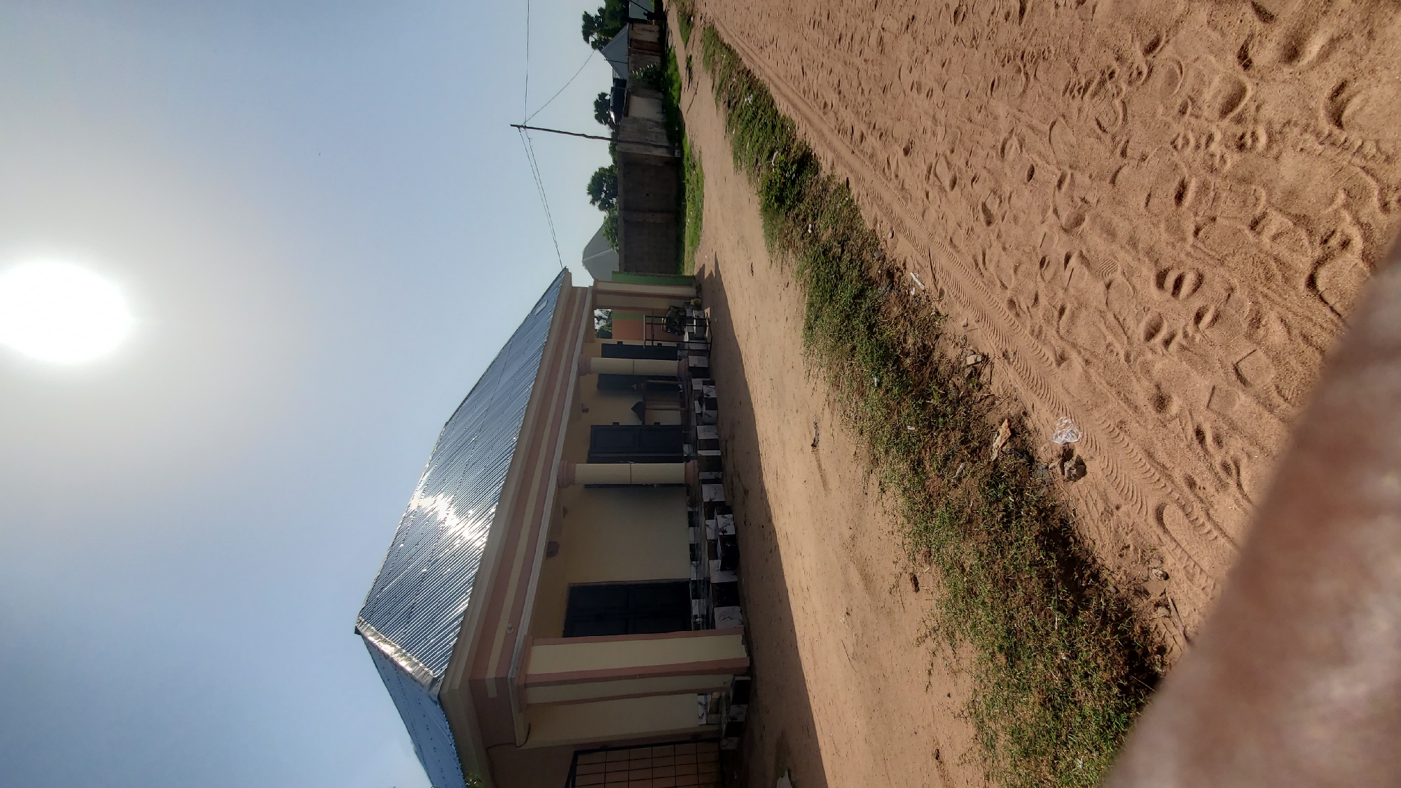 A one-story, light yellow building with a corrugated metal roof and white and pink trim sits on the left side of a dusty, unpaved road in Jalingo, Nigeria. The building has a covered porch supported by four cylindrical columns, with dark-colored doors and windows. The ground in front of the building is paved with black and white checkered tiles. A fence with vertical bars is visible to the left of the building entrance. The road leading away from the building is sandy and marked with tire tracks and footprints. Vegetation, including grass and some small trees, grows along the edge of the road and beyond a low wall in the background. The sky is clear and bright blue with a prominent sun in the upper right portion of the image. The scene appears to be a residential or commercial property in a rural or semi-urban setting.