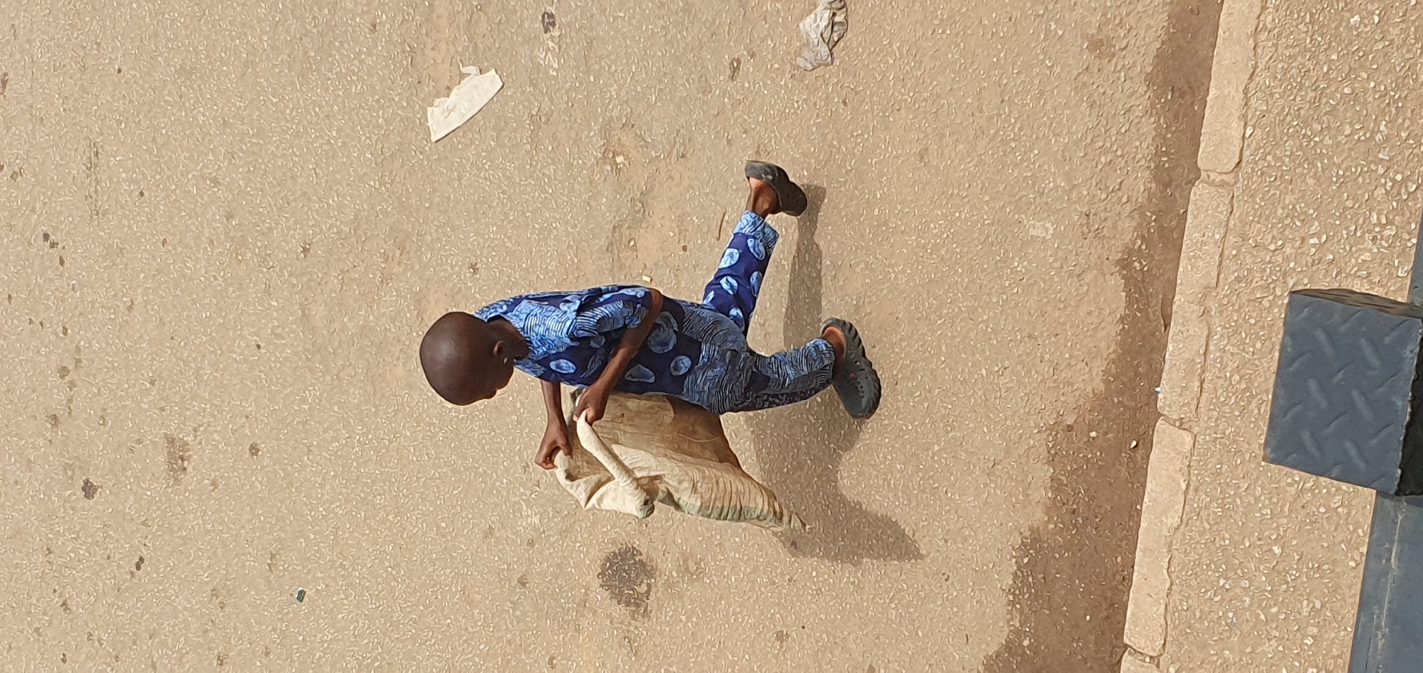 In Bukuru, Nigeria, a young boy is captured in a high-angle shot, walking on a paved surface. He is wearing a blue patterned outfit, consisting of a shirt and trousers, and dark clogs on his feet. The boy is carrying a large, light-colored sack over his left shoulder, holding its strap with both hands. His body is angled to the right, with his left leg forward in a walking stride, casting a shadow on the ground. The environment is a dusty, light-brown paved area with scattered debris, including a crumpled piece of white paper to the right of the boy. A raised curb with a light-colored stone border is visible at the bottom edge of the frame, with a dark metal object positioned in the foreground.
