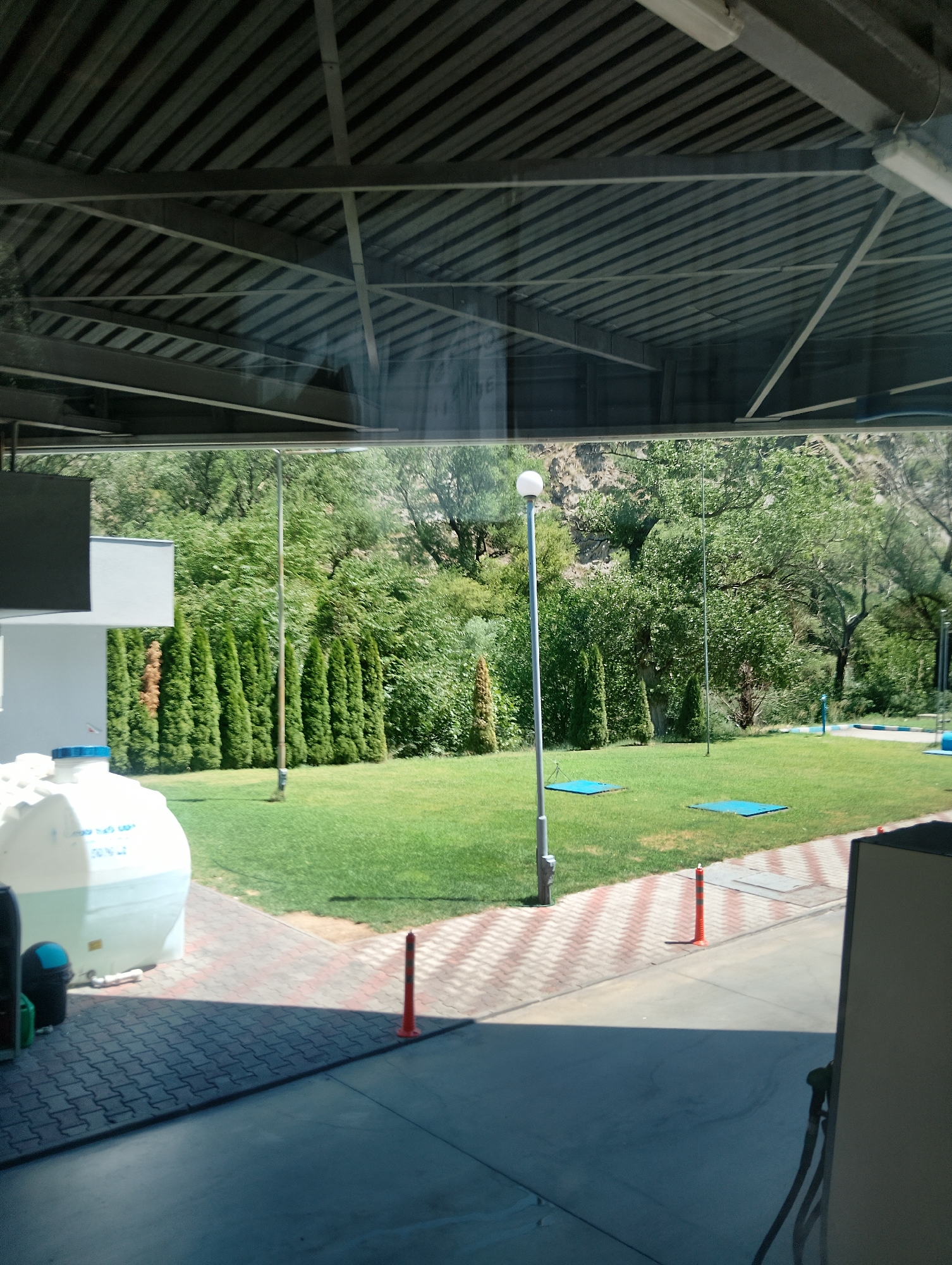 The image depicts an outdoor scene, likely a park or recreational area adjacent to a covered structure, viewed through a reflective surface. The foreground shows a paved walkway with a pattern of red and white rectangular tiles, and a darker grey concrete area. A large white plastic water tank with blue markings is positioned on the left side of the walkway. To the left of the tank, a black trash receptacle with a blue lid is visible. Two orange traffic cones stand on the paved area, one near the water tank and another further down.
Beyond the paved area is a well-maintained green lawn. Several tall, slender evergreen trees are planted in a line along the edge of the lawn, behind which are dense green trees and shrubs forming a backdrop. A tall, thin lamppost with a spherical light fixture stands on the lawn. Two blue rectangular mats are placed on the grass in front of the lamppost. In the middle ground, the terrain slopes upwards, covered with vegetation. The background shows a rocky hillside with trees. A person can be seen in the far right of the image, in the distance, standing on the lawn.
The upper part of the image is dominated by a reflected view of a corrugated metal roof structure with visible beams and supports. The reflection also shows some of the surroundings, including trees and the sky. The overall impression is of a well-kept outdoor space, possibly at a service station or a community area, given the presence of the water tank and traffic cones, situated in a location with a natural, vegetated environment.