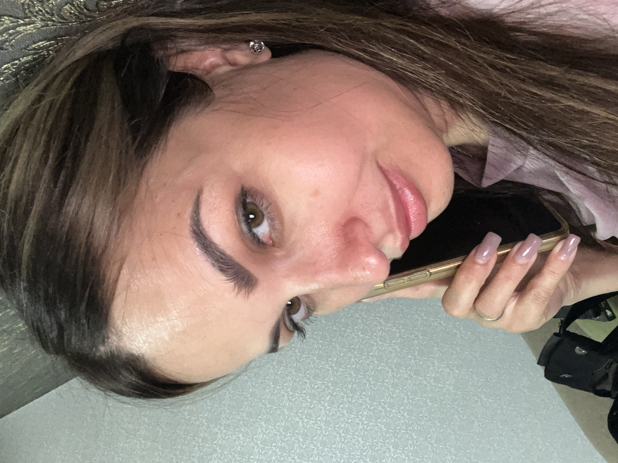 A woman with long, brown hair, wearing a pink shirt and a silver ring on her left ring finger, is holding a gold smartphone to her right ear and speaking on it. She is positioned at a slight angle, looking directly at the camera with her head tilted. A diamond stud earring is visible in her right ear. The background consists of a textured, light blue wall on the left and dark brown hair on the right.