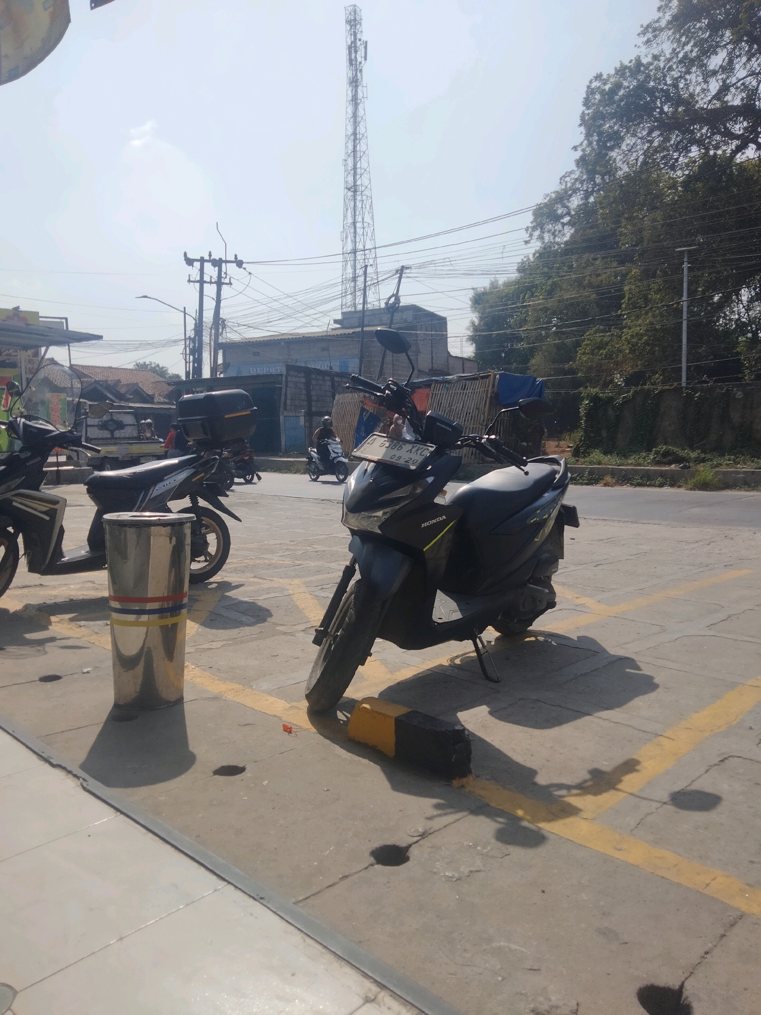 The image depicts a street scene in Pisangan Jaya, Indonesia. In the foreground, several motorbikes are parked on a paved surface marked with yellow lines. A cylindrical metal trash bin with red and yellow bands stands near the center. To the right, a black and yellow parking bumper is visible, casting a shadow. Beyond the parked motorbikes, a road runs horizontally, with a lone motorbike rider on it in the middle ground. Buildings line the street, including a structure with the word "DEPOT" visible on its facade. Overhead, a prominent telecommunications tower dominates the sky, alongside numerous electrical wires. Trees with dense foliage are visible on the right side of the image, and a pale, hazy sky suggests daylight.