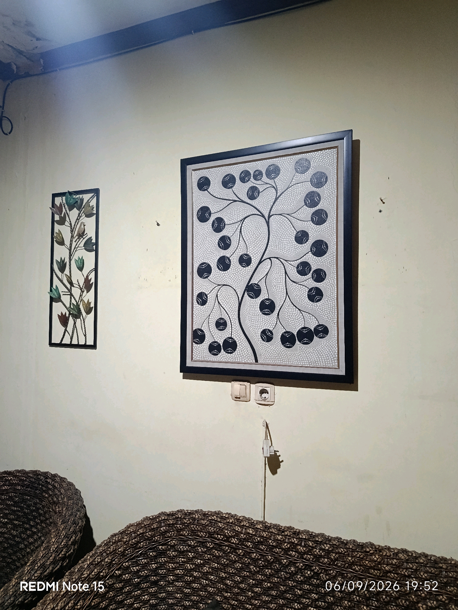 The image depicts a dimly lit interior scene, likely a living room, with furniture and wall art. Two woven, dark brown chairs or a sofa occupy the foreground, their textured surfaces illuminated by a light source from the left. A wall with a textured, cream-colored surface dominates the background.
Mounted on the wall are two pieces of art. To the left, a rectangular framed artwork features a metal sculpture of a bird perched on a branch with stylized leaves and flowers in muted green, blue, and red. To the right, a larger square framed artwork displays a black and white graphic of a tree with circular fruits or leaves, rendered in a dot-work style on a textured background. Below this artwork, a white electrical outlet with two sockets and a light switch are visible. A white plug is partially inserted into one of the sockets, with a thin yellow wire extending downwards.
In the upper left corner of the image, a dark, possibly smoky or dusty band is visible along the ceiling line, with some electrical wiring. A timestamp and "REDMI Note 15" are overlaid in white text at the bottom left of the image, and a date and time stamp "06/09/2026 19:52" is overlaid at the bottom right. The scene appears to be domestic, devoid of human presence. No specific landmarks for Keboananom, Indonesia, are identifiable within the frame.