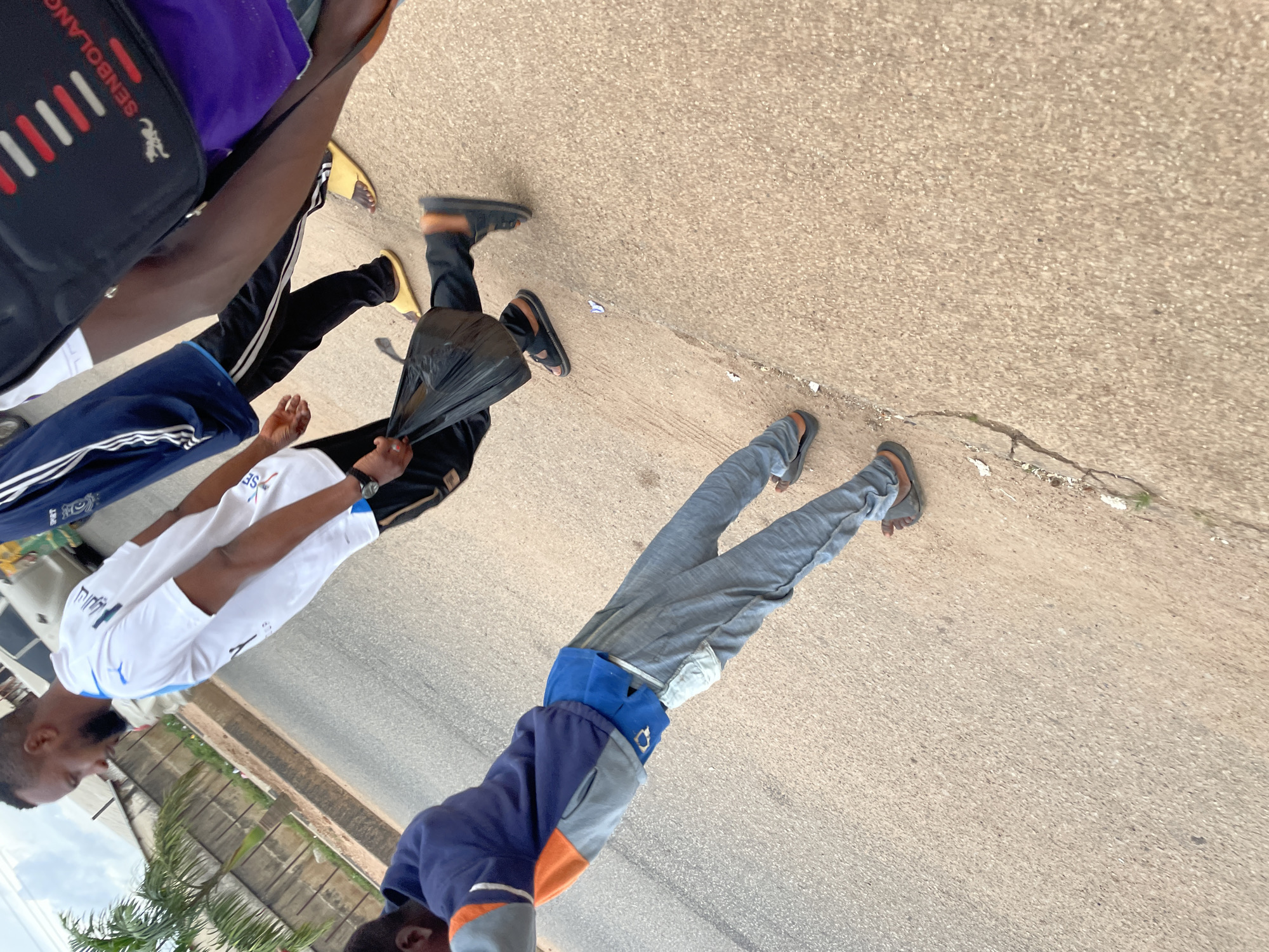 The image depicts a street scene in Barakin Lerawi, Nigeria. Three individuals are visible, walking on an asphalt road.
The person closest to the foreground is wearing a blue and orange jacket over a blue shirt and gray sweatpants. They are also wearing gray sandals.
The second person, to the left and slightly behind the first, is wearing a white t-shirt with blue accents and black shorts. They have a black plastic bag in their right hand and are wearing black sandals.
The third person, further in the background and to the right, is wearing a blue and black tracksuit and yellow sandals. A portion of a black bag with red and white stripes is visible on their left shoulder.
The environment includes an asphalt road with some cracks, a concrete barrier or wall on the left, and a palm tree. A white vehicle is partially visible in the background. The sky is cloudy.