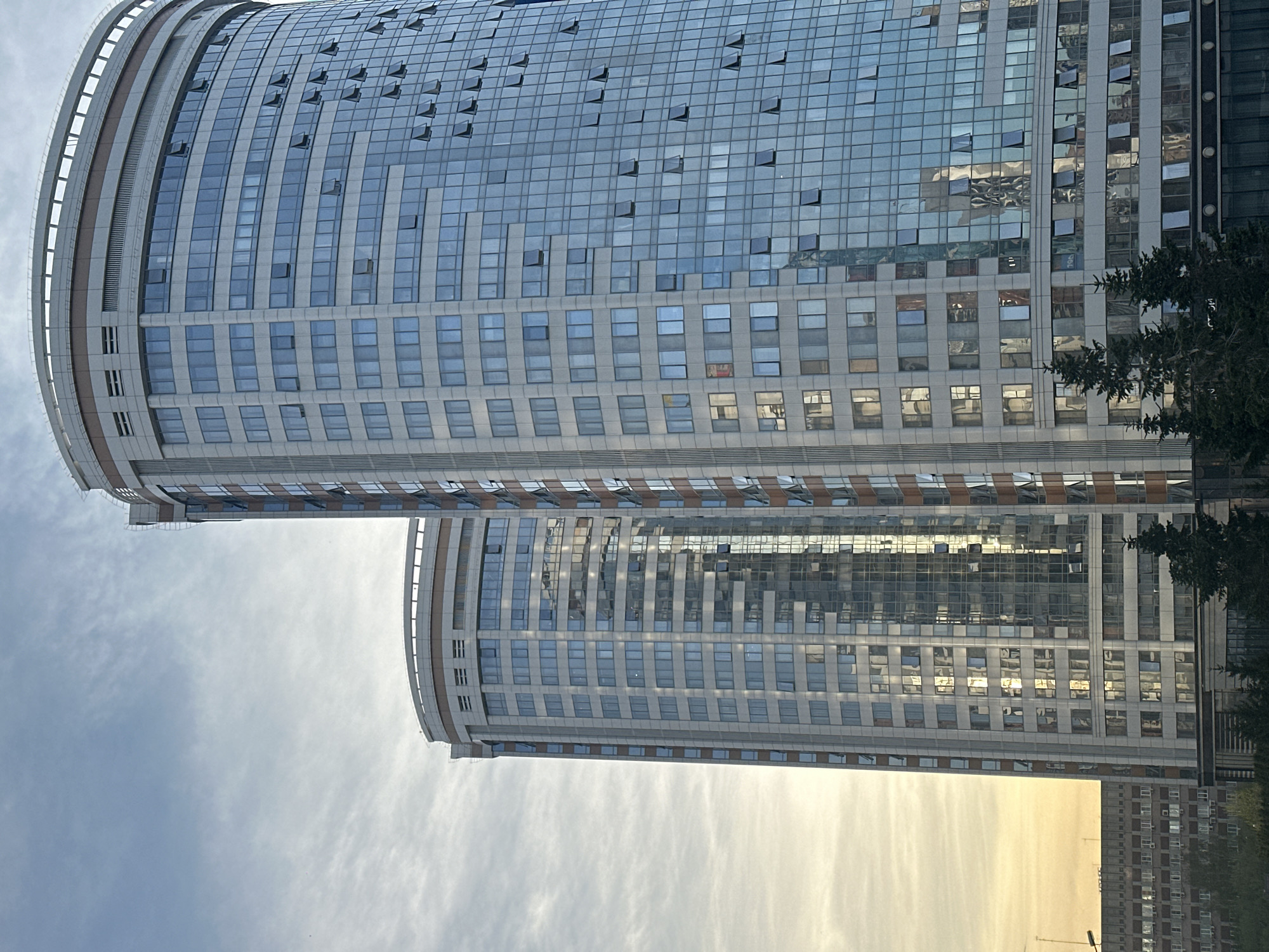 The image depicts two tall, modern, curved buildings with numerous windows in Novosibirsk, Russia. The buildings have a similar design with a rounded facade. Many windows appear open. Reflections of the sky and other buildings are visible on the glass surfaces. Trees are visible in the lower right corner. The sky is cloudy.