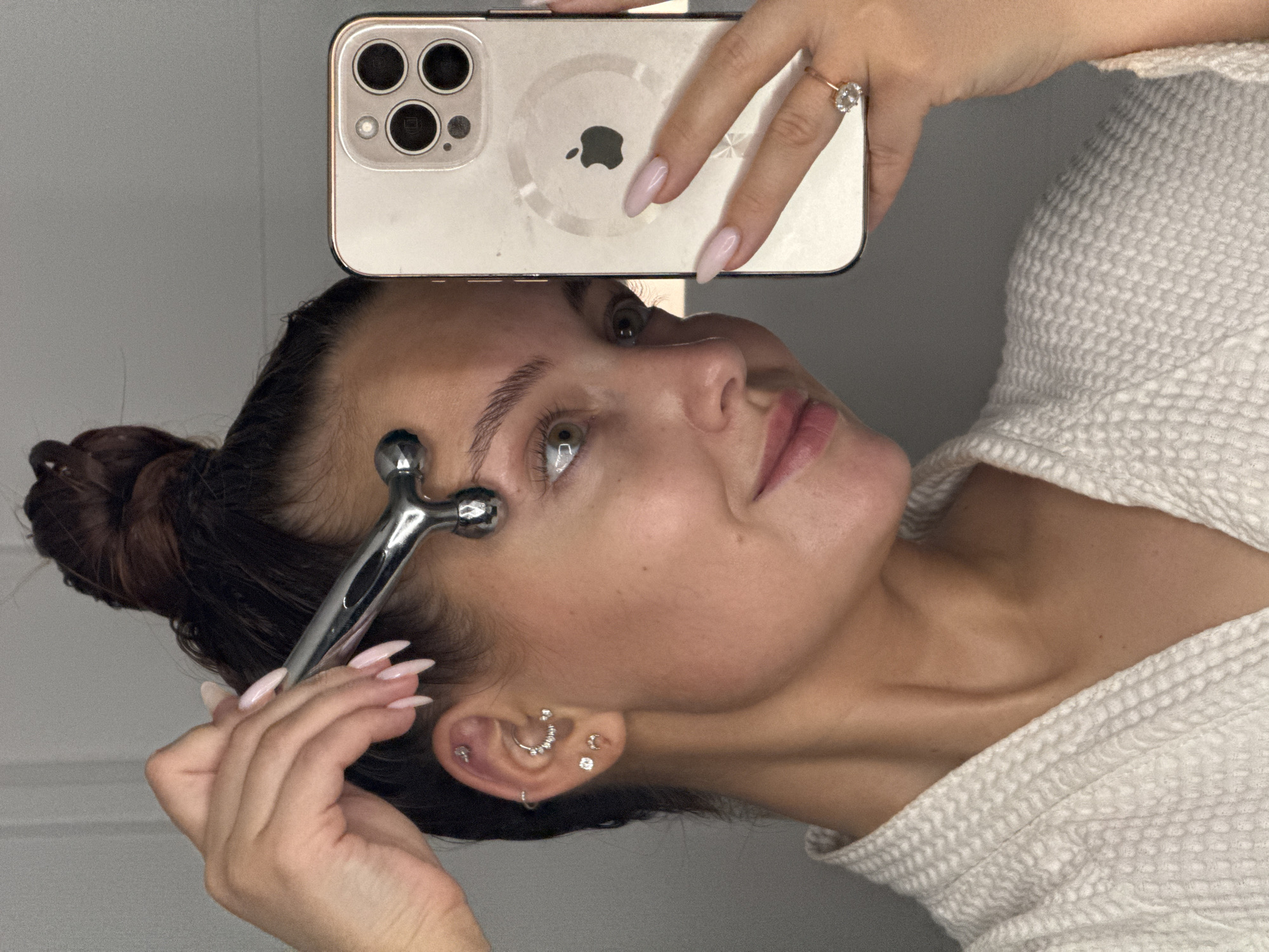 A woman is holding a smartphone above her forehead, using it to take a selfie. She is also holding a silver facial roller to her face. She has her hair pulled back in a bun and is wearing a light-colored robe. Her nails are painted light pink, and she has multiple earrings in her left ear. The background appears to be a plain gray wall. There is no information in the image to indicate this photo was taken in Chaikovsky, Russia.