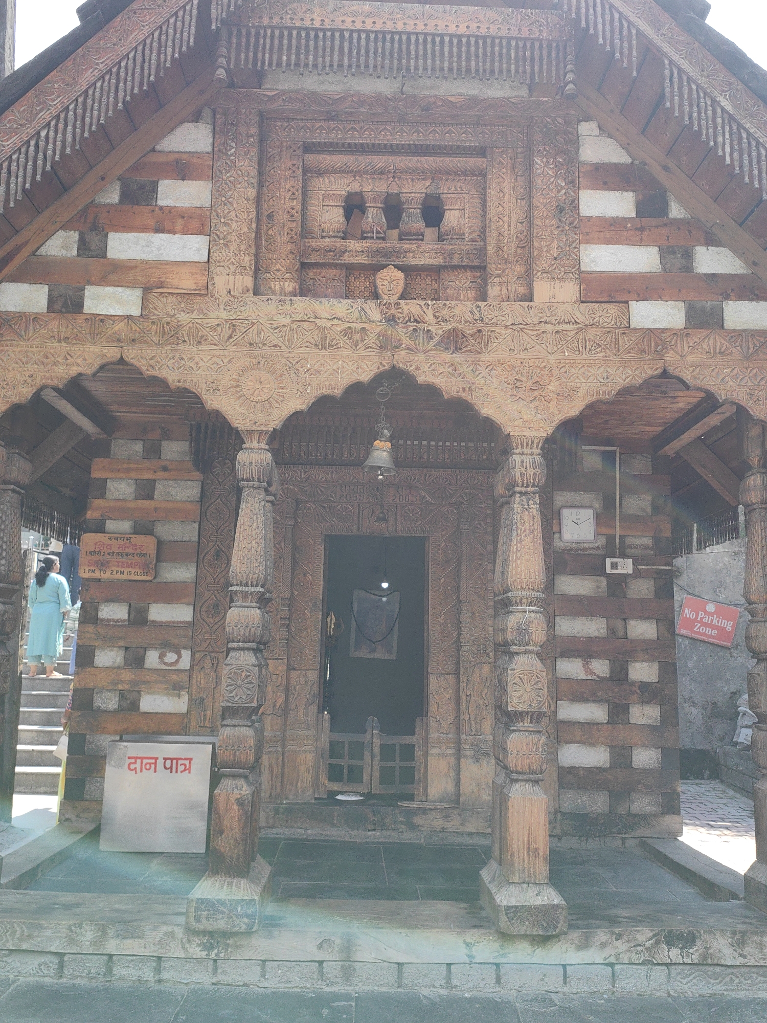 The image depicts the entrance to a wooden temple in Vashisht, India. The temple features intricate wood carvings and is constructed with a combination of wood and stone. A bell hangs above the entrance. To the left, a woman in a light blue dress is ascending a set of stone steps. A sign with Hindi text is visible on the wall to the left of the entrance, and another sign in Hindi reading "दान पात्र" (Daan Patra, meaning donation box) is located on the lower level. To the right of the entrance, a clock is mounted on the wall, and a "No Parking Zone" sign is also present. The overall scene suggests a cultural and religious site bathed in sunlight.
