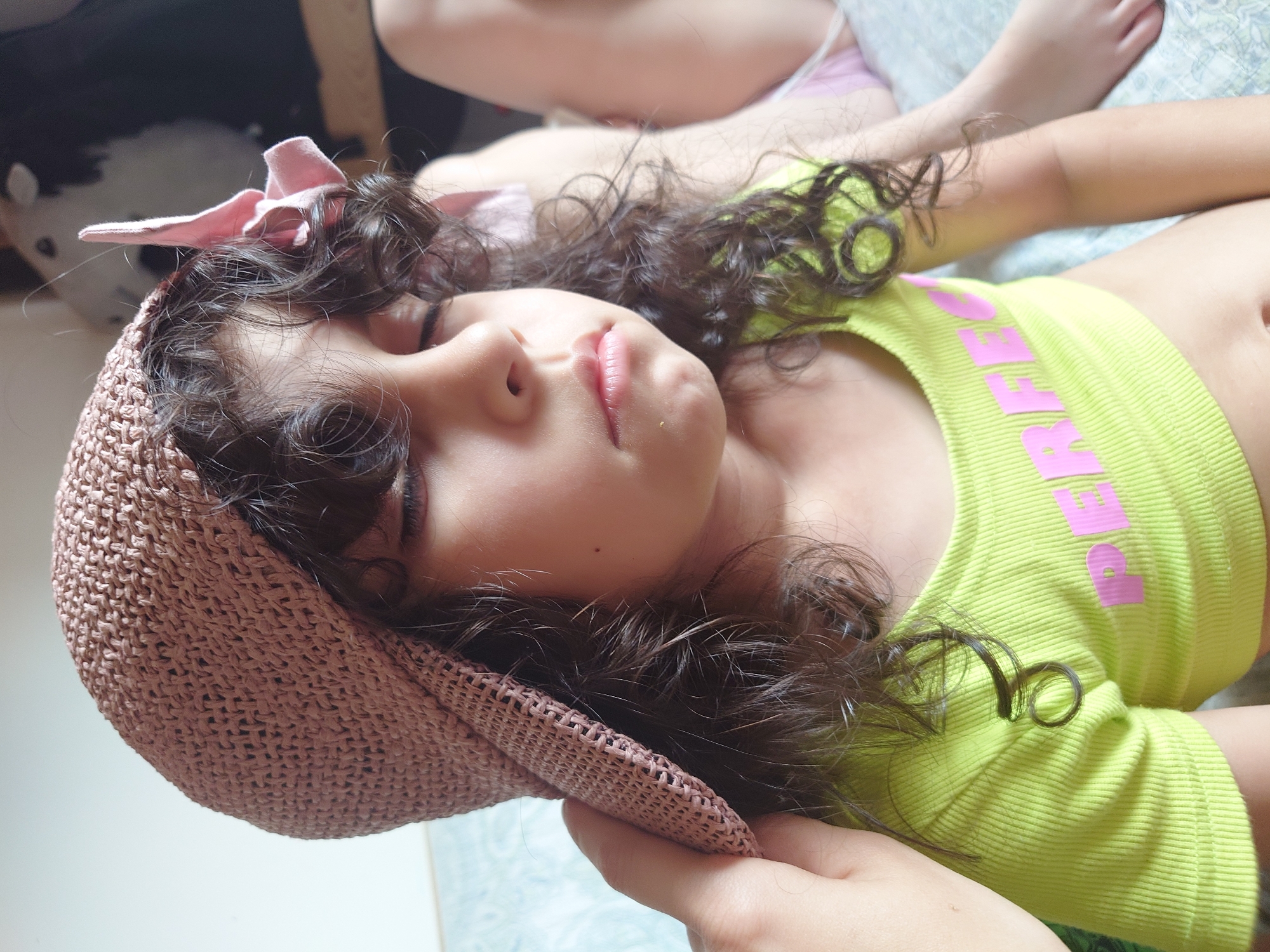 A young child with curly dark hair, wearing a light pink straw hat with a pink bow and a lime green ribbed crop top with pink lettering, is shown from the chest up, with their eyes closed. Their left hand is visible near the hat. In the background, another child's legs are visible, and the scene appears to be indoors on a patterned surface, possibly a bed or couch. The location is the city of Kommunarka, Russia.