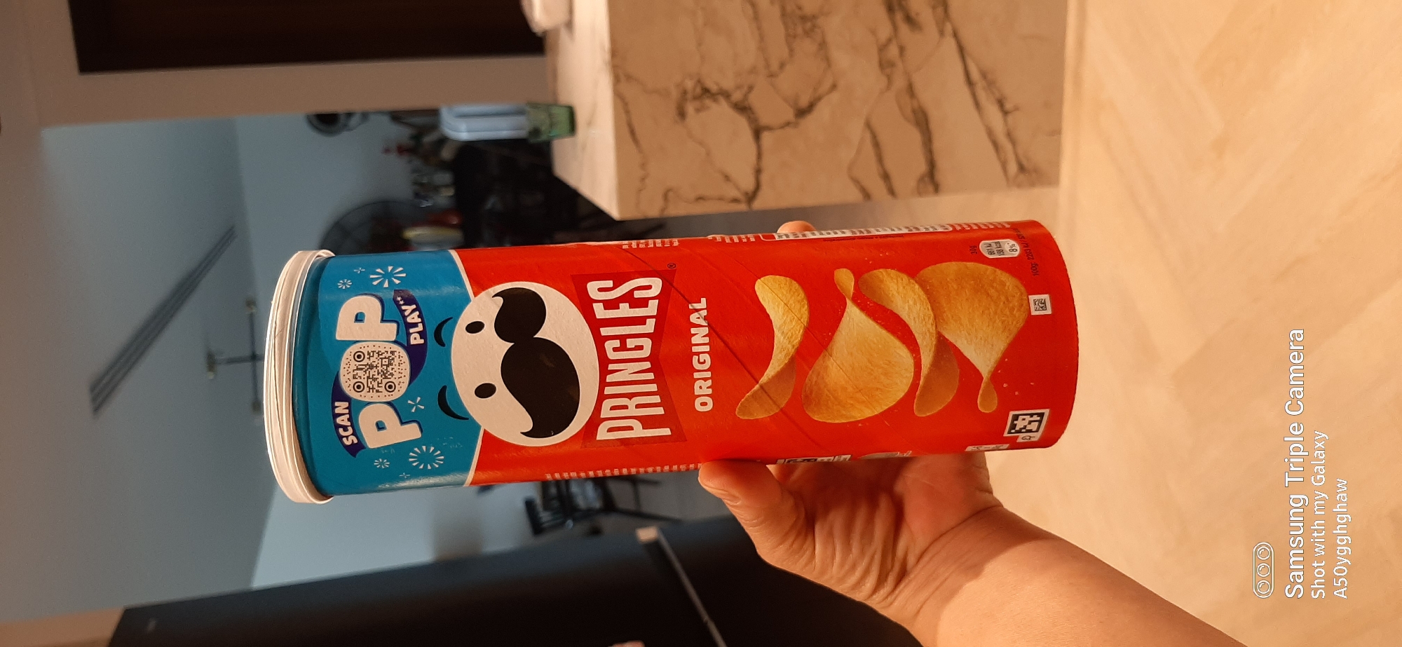 An image captured in an indoor setting, likely a kitchen or dining area in Dubai, United Arab Emirates, features a person's hand holding a Pringles "Original" flavor can. The hand, with visible lines and skin texture, grasps the red and blue cylindrical container.
The Pringles can is prominently displayed, oriented horizontally. The red portion features the "PRINGLES ORIGINAL" text, along with the iconic Mr. Pringle's face. Below the text, several crispy, golden potato chips are illustrated. Small nutritional information is visible on the red section, including "309" and partially "100g: 22". The blue section of the can displays the text "SCAN" and "POP PLAY" next to a QR code, surrounded by white starbursts.
In the background, a light-colored, possibly marble-patterned countertop is visible, suggesting a modern interior. The lighting is bright and even, indicating a daytime shot or well-lit interior. The overall scene is clean and focused on the product. A watermark in the bottom right corner reads "Samsung Triple Camera Shot with my Galaxy A50ygghghaw", indicating the device used to capture the image.