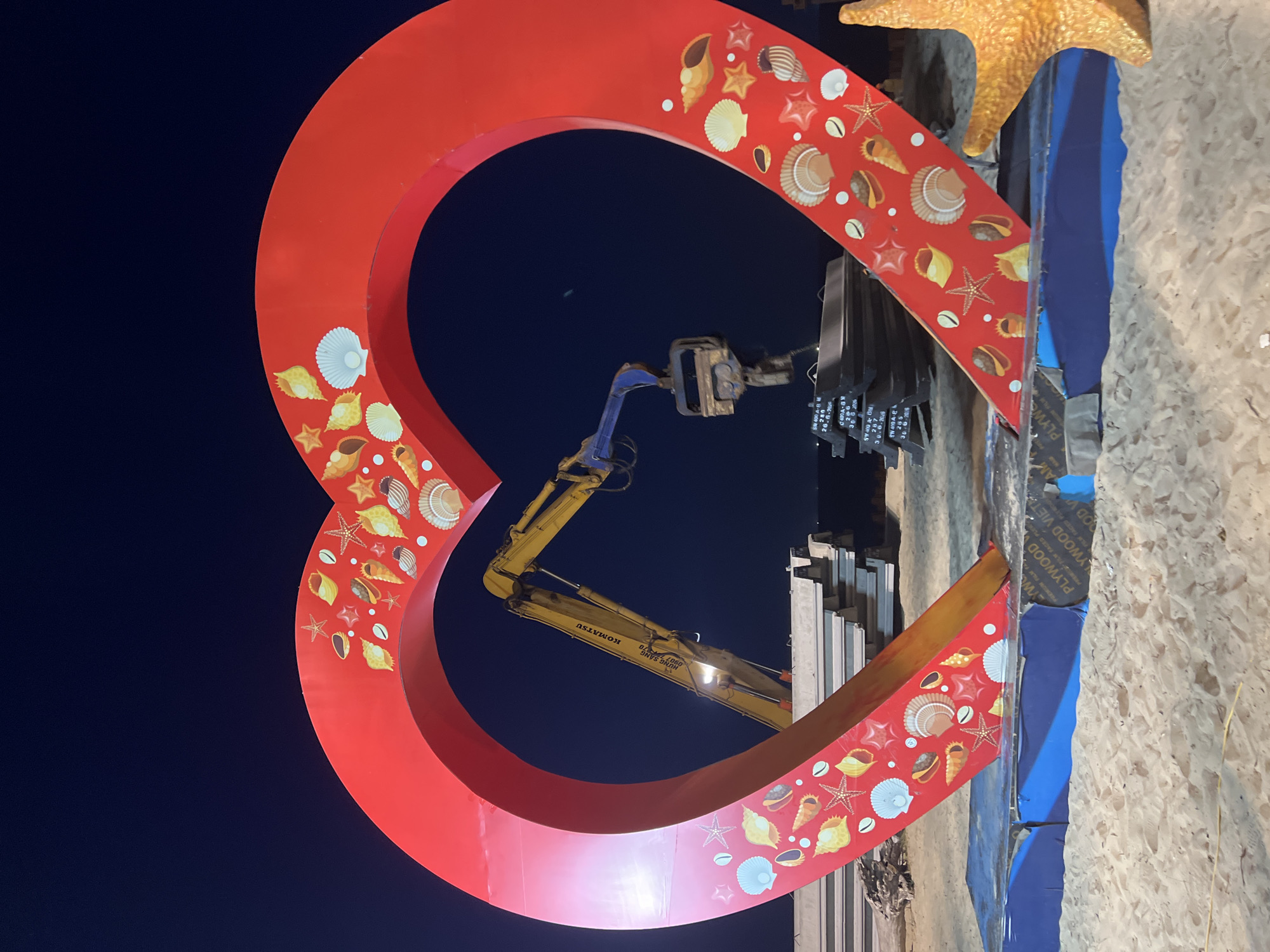 This image captures a large, red heart-shaped structure decorated with various seashells and starfish, situated on a sandy beach at night. The structure is illuminated from within, casting a bright glow. Behind the heart, a yellow Komatsu excavator is visible, its arm extended and holding a clamp. In the background, stacks of what appear to be construction materials or steel beams are arranged. The scene is located in Hoi An, Vietnam.