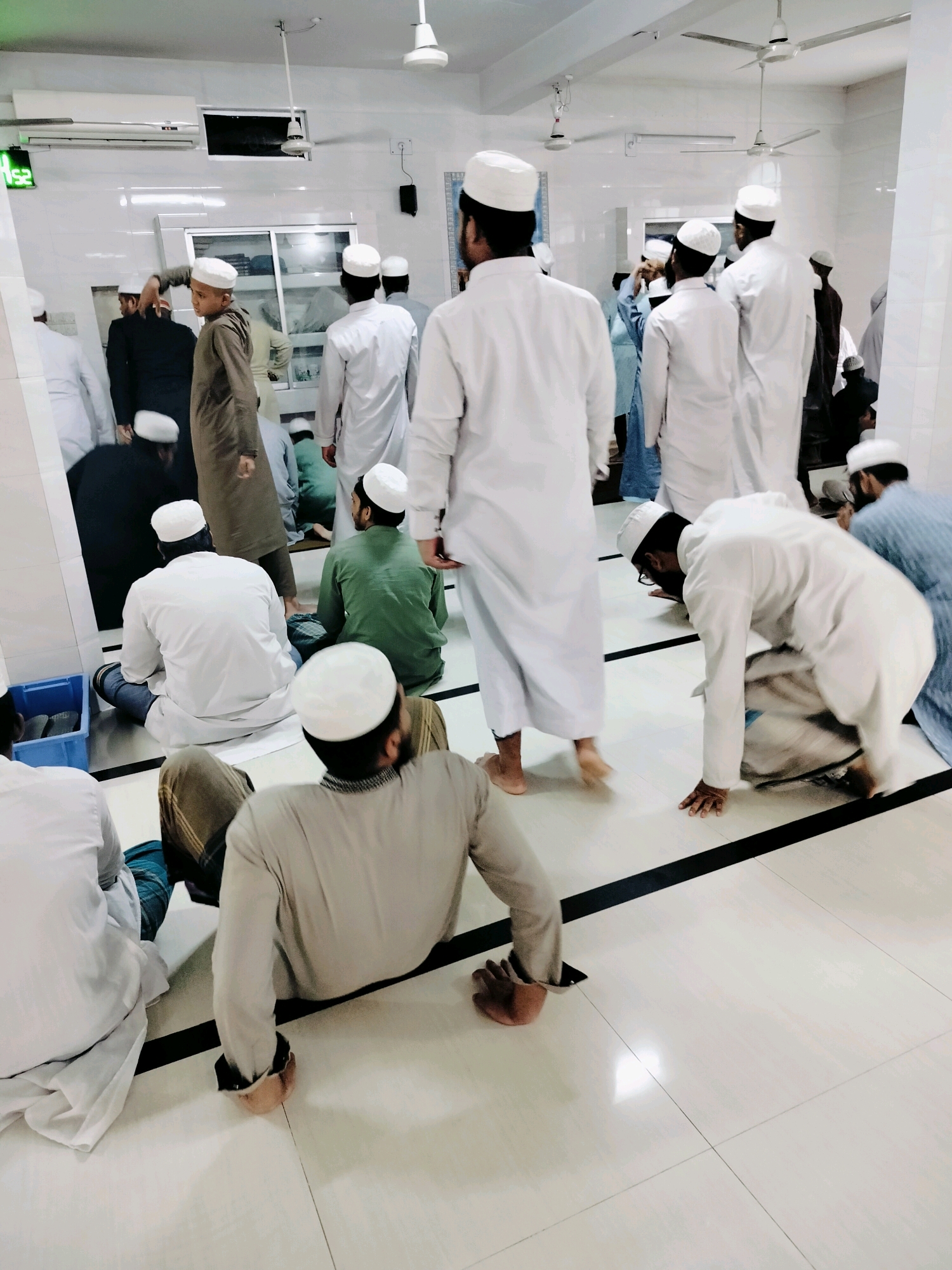 A group of men in white religious attire are gathered in a mosque. Many are wearing white kufis, and some are engaged in prayer. One man is bowing down on the floor, with his hands on the ground. Others are sitting or standing, with some observing. The floor is tiled with a grid pattern, and there are white walls and ceiling lights. A digital clock on the wall shows "12:52". A blue bin is visible in the lower left corner. The scene appears to be in Dhaka, Bangladesh.