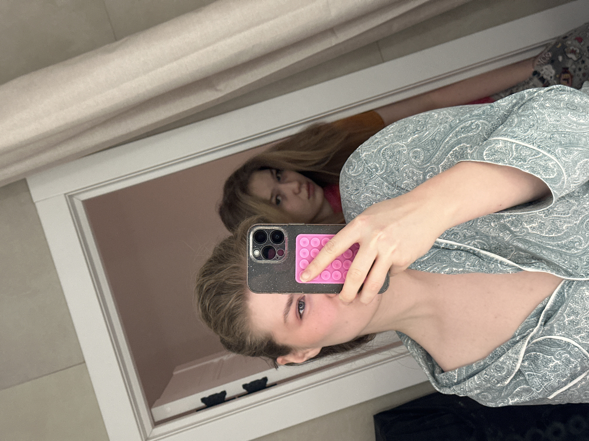 A woman is taking a selfie in a mirror. She has blonde hair and is wearing a light blue paisley patterned pajama top. The reflection in the mirror shows another woman with long blonde hair wearing a brown top, partially obscured by the selfie-taker. The phone case on the selfie-taker's phone is dark gray with a pink pop-out grip. The background includes a beige wall or curtain and a white doorframe. Based on visual information, it is not possible to confirm the location in Kazan, Russia.