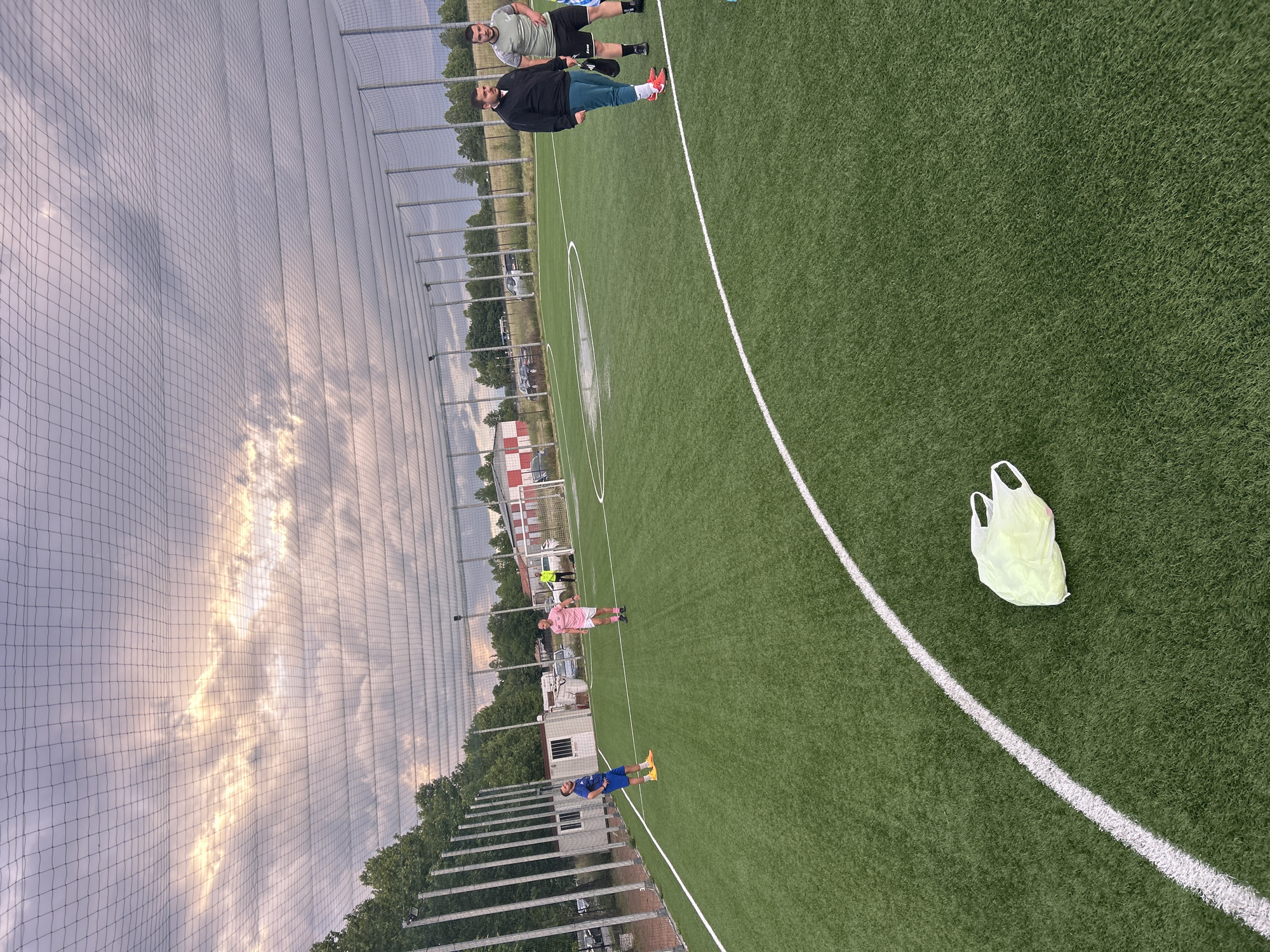 The image depicts an artificial turf football pitch, likely in Kazanlak, Bulgaria, during the late afternoon or early evening. The sky, visible through a tall protective netting, is partly cloudy with sunlight breaking through, casting a soft glow.
Several men are standing on the vibrant green pitch. In the mid-ground, a man wearing a black jacket, dark green pants, and red and white sports shoes stands with another man in a grey t-shirt and black shorts. Further down the pitch, another man in a pink t-shirt and black shorts is visible, along with a man in a blue football kit and yellow shoes. The men appear to be gathered casually, possibly before or after a game.
A light yellow plastic bag rests on the artificial turf near a prominent white line that stretches across the foreground. The pitch features standard white markings, including a portion of the center circle. The playing area is enclosed by a tall, dark mesh netting that separates it from the surrounding environment. Beyond the netting, there are green trees, several buildings (one with distinctive red and white checkered patterns), and parked cars, indicating a sports facility or complex. No visible text is present in the image.