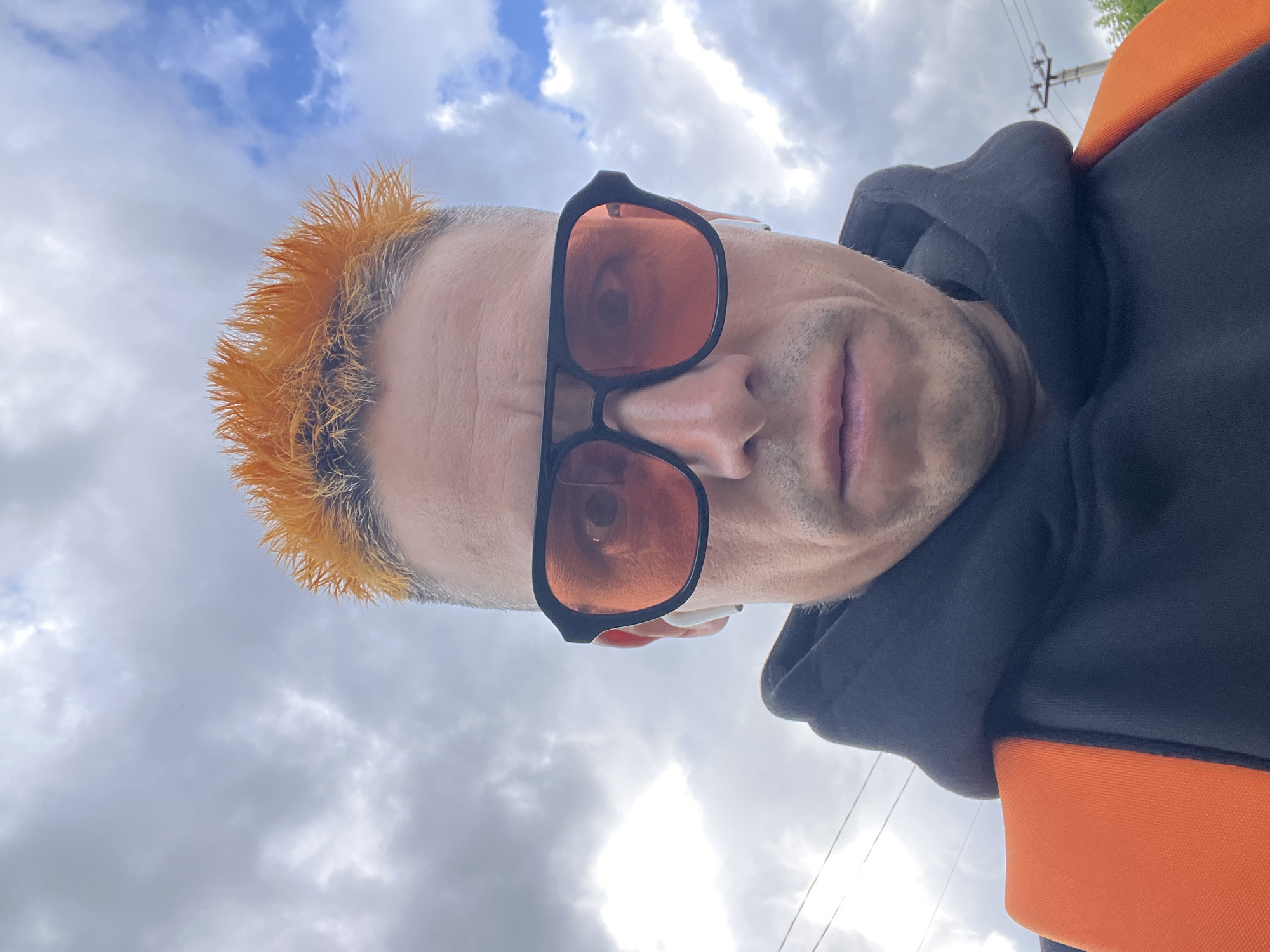 A close-up, vertically oriented selfie shows a person's face and upper body against a cloudy sky. The person has bright orange, spiky hair. They are wearing large, black-framed sunglasses with orange lenses that reflect the sky. A dark hoodie with an orange trim is visible. The person appears to be looking towards the camera. The sky is filled with white, fluffy clouds, and there are hints of electrical wires in the upper right portion of the frame, suggesting an outdoor, possibly urban or semi-urban setting. The lighting suggests it is daytime, but the cloud cover makes it difficult to determine the exact time. No text is visible in the image. The overall mood is casual and the pose is informal. The image was likely taken in Chaikovsky, Russia.