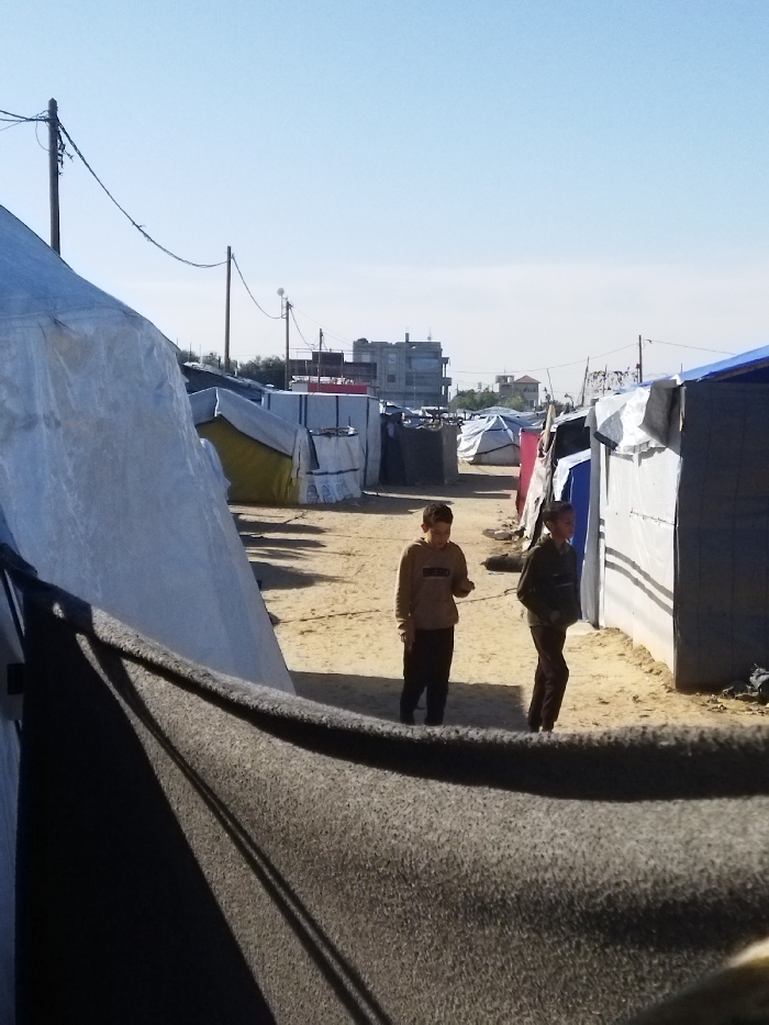 The image depicts an informal settlement or refugee camp located in Khan Yunis, Palestinian Territory, under a clear, bright blue sky. In the immediate foreground, a dark grey, coarse-textured fabric or blanket is partially visible, extending across the lower portion of the frame and casting a shadow.
The central part of the scene features a wide, unpaved dirt path, light brown in color, that extends into the distance. This path is flanked on both sides by numerous makeshift shelters and tents. These structures are constructed from various materials, primarily white, light blue, and grey tarpaulins and fabrics, showing signs of piecemeal assembly and varying conditions. A large white tent-like structure is prominent on the left side of the foreground.
Two young boys are present on the dirt path. The boy on the left wears a light brown hooded sweatshirt with a dark graphic and dark pants; he is looking downwards and appears to be holding a small object in his right hand. The boy on the right wears a dark, possibly striped, long-sleeved top and dark pants; he is looking towards the boy on the left with a slight smile.
In the mid-ground and background, more shelters line the path. Further in the distance, several multi-story concrete buildings are visible, indicating the proximity of a more urban area. Utility poles with overhead wires traverse the scene, extending into the background. Shadows are distinctly cast on the ground and structures, suggesting direct sunlight.