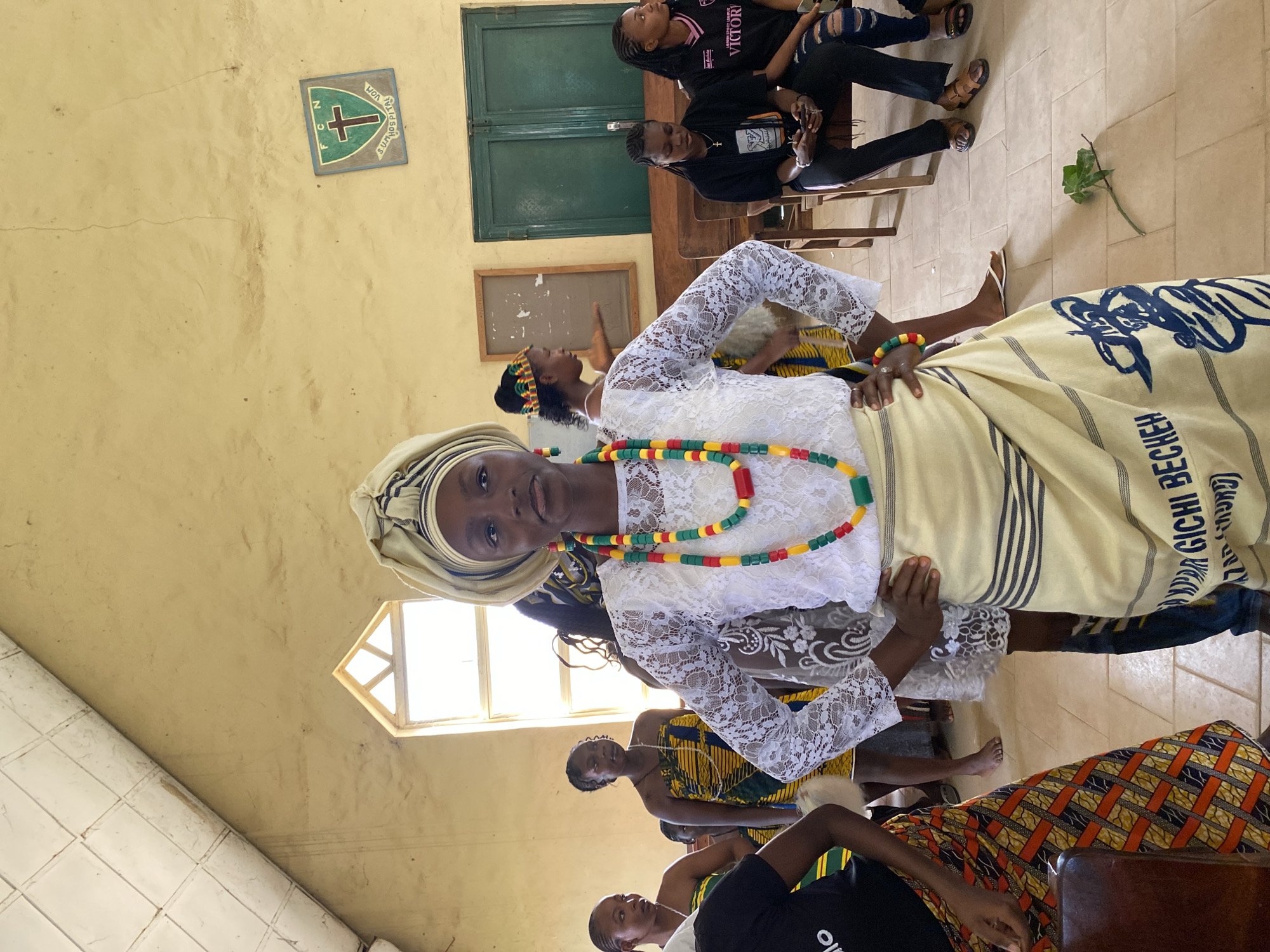 In an indoor setting with pale yellow walls, a group of young Black women are gathered, possibly for a cultural event or performance. The primary subject in the foreground, a young woman, is dressed in traditional attire. She wears a white lace top and pants, a beige headscarf, and a beaded necklace in red, yellow, and green. A cream-colored cloth with dark stripes and blue text is draped over her. Behind her, other women are seen, some wearing vibrant patterned clothing, including a woman in a yellow, green, and blue kente-like fabric and another in a black t-shirt. One woman in the background is wearing a black t-shirt with "VICTORY" printed on it, and ripped blue jeans.
The setting appears to be a room with wooden benches and a window with a geometric frame. On the wall, a green and blue shield with a cross and the letters "F C N" is visible. Another framed object is present on the wall. The overall atmosphere seems to be one of community and celebration, with the women appearing engaged and attentive, and the foreground subject smiling. The lighting suggests it is daytime, possibly late morning or afternoon, with natural light coming from the window. The floor is tiled. The text visible on the cream cloth reads "GICHI BECKEN".